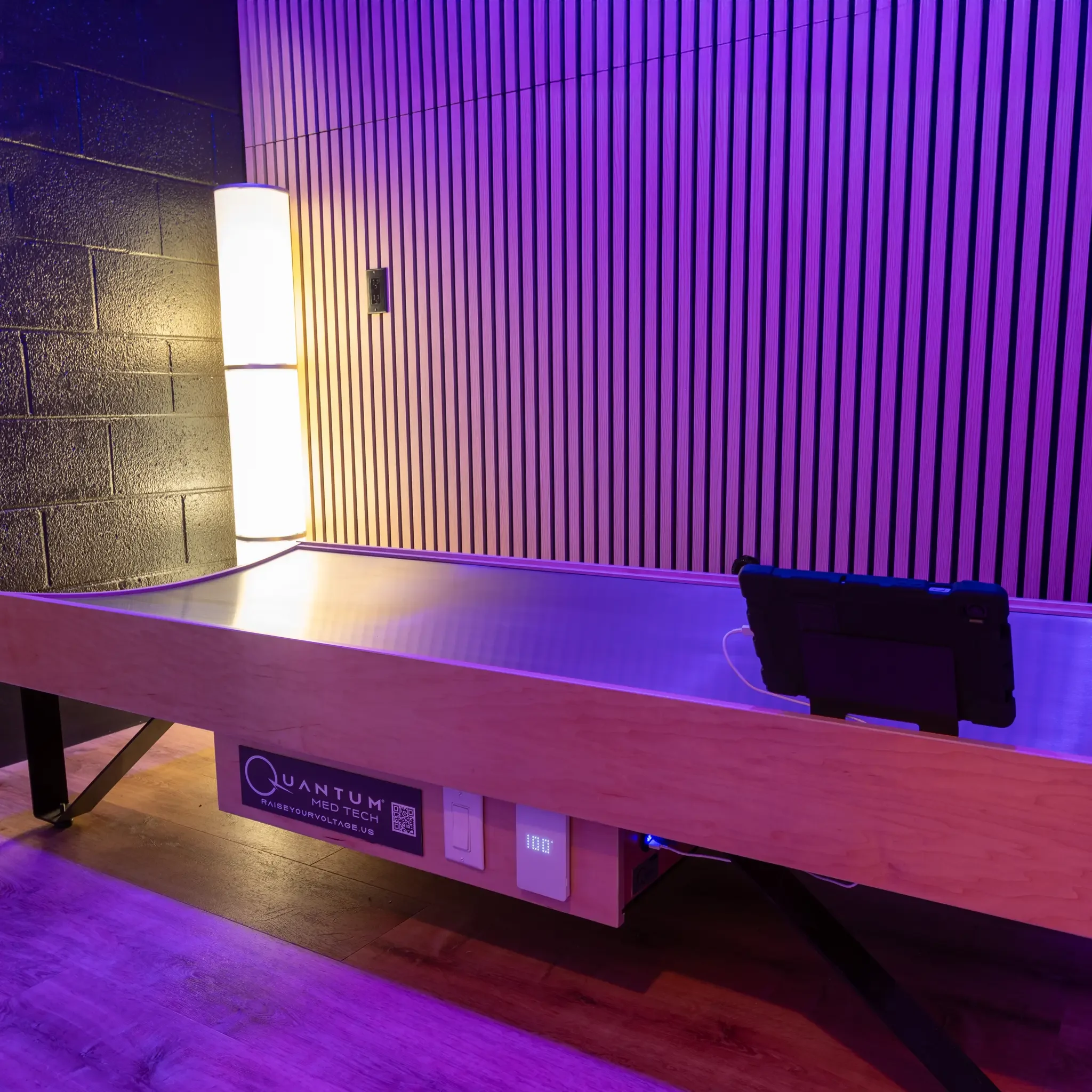 Interior of a room with a purple-lit slatted wall, a white wall sconce, and a wooden counter with a QR code sign for Quantum bed.