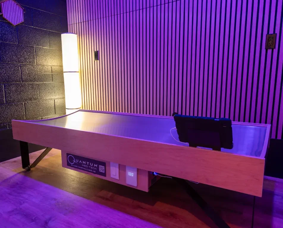 A Quantum bed in a room with purple and pink lighting, featuring a wooden exterior, black legs, and control panels on the front, with a chair nearby.