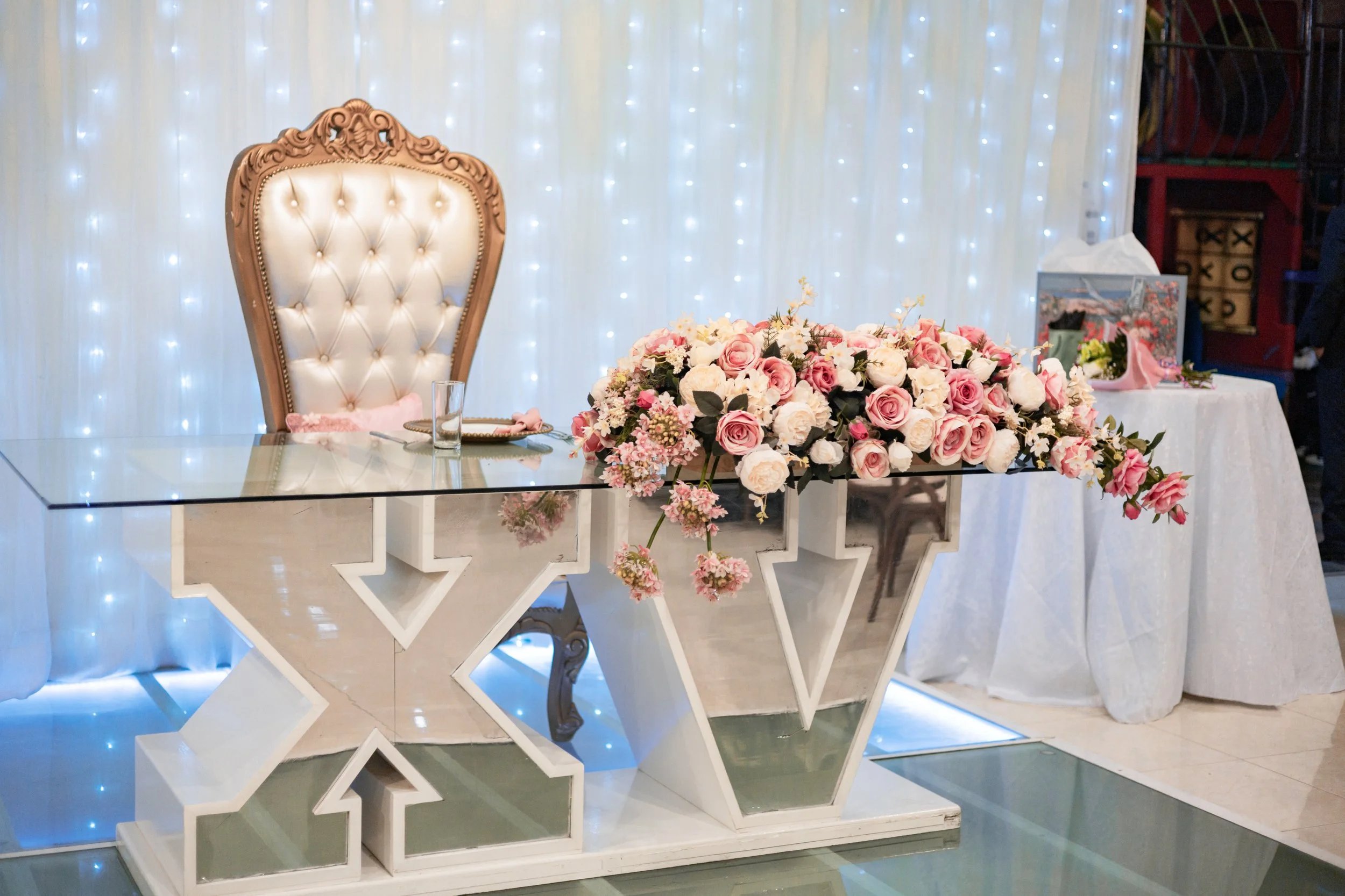 A wedding reception setup with a decorated table, a large ornate chair, and a floral arrangement of pink and white roses on the table.