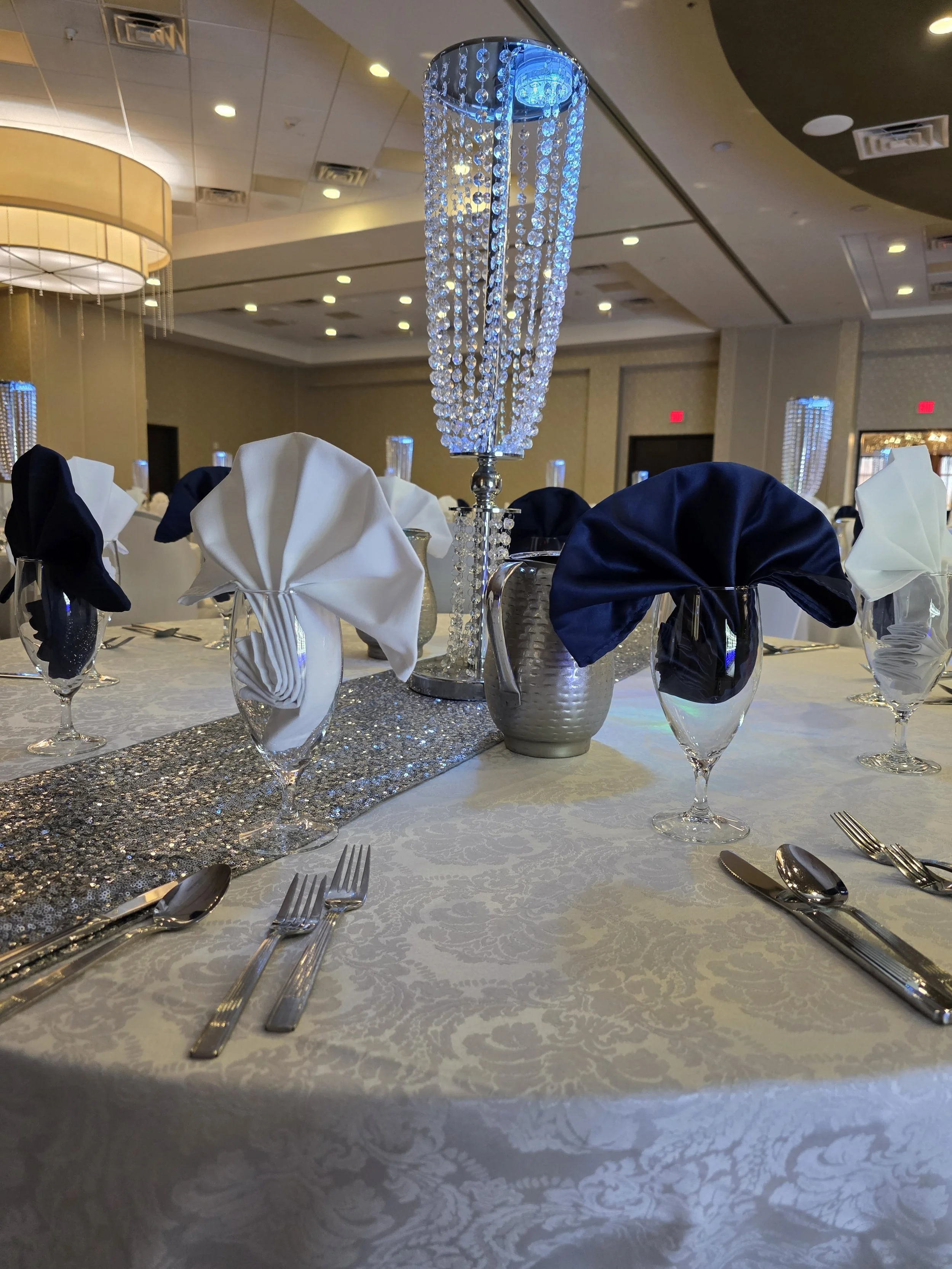 A decorated banquet table set for an event, with black and white napkins folded in a fan shape, silverware, wine glasses, a silver vase, and a tall crystal chandelier centerpiece.
