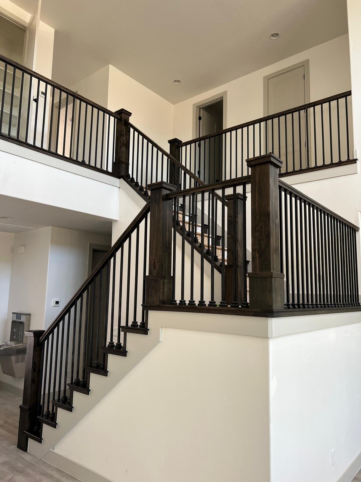 Interior view of a two-story house staircase with dark wood handrails and balusters, beige walls, and two doors on the upper level.