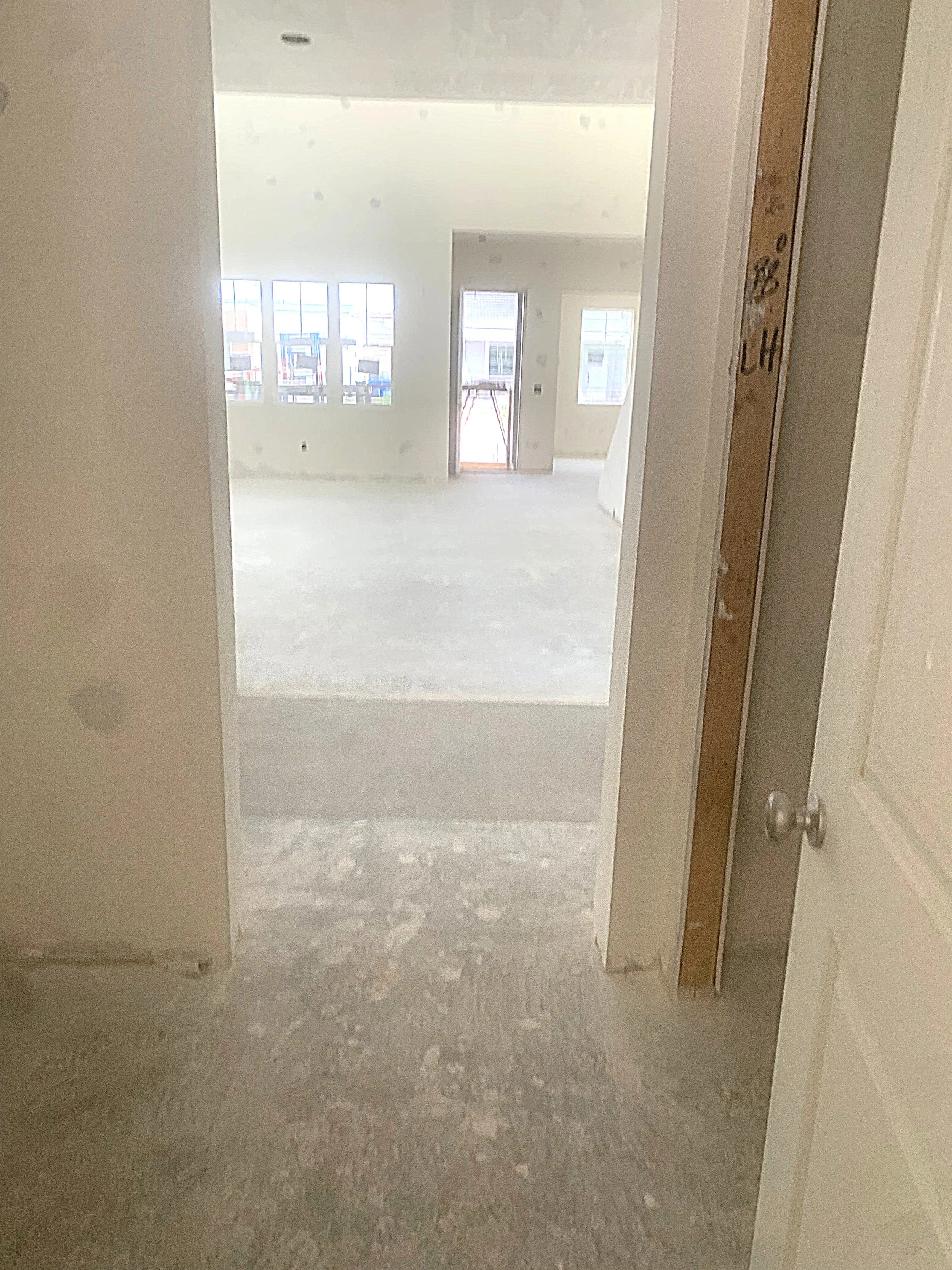 An interior view of an unfinished room in a building under construction, showing a doorway leading into a bright room with multiple windows and a balcony door.