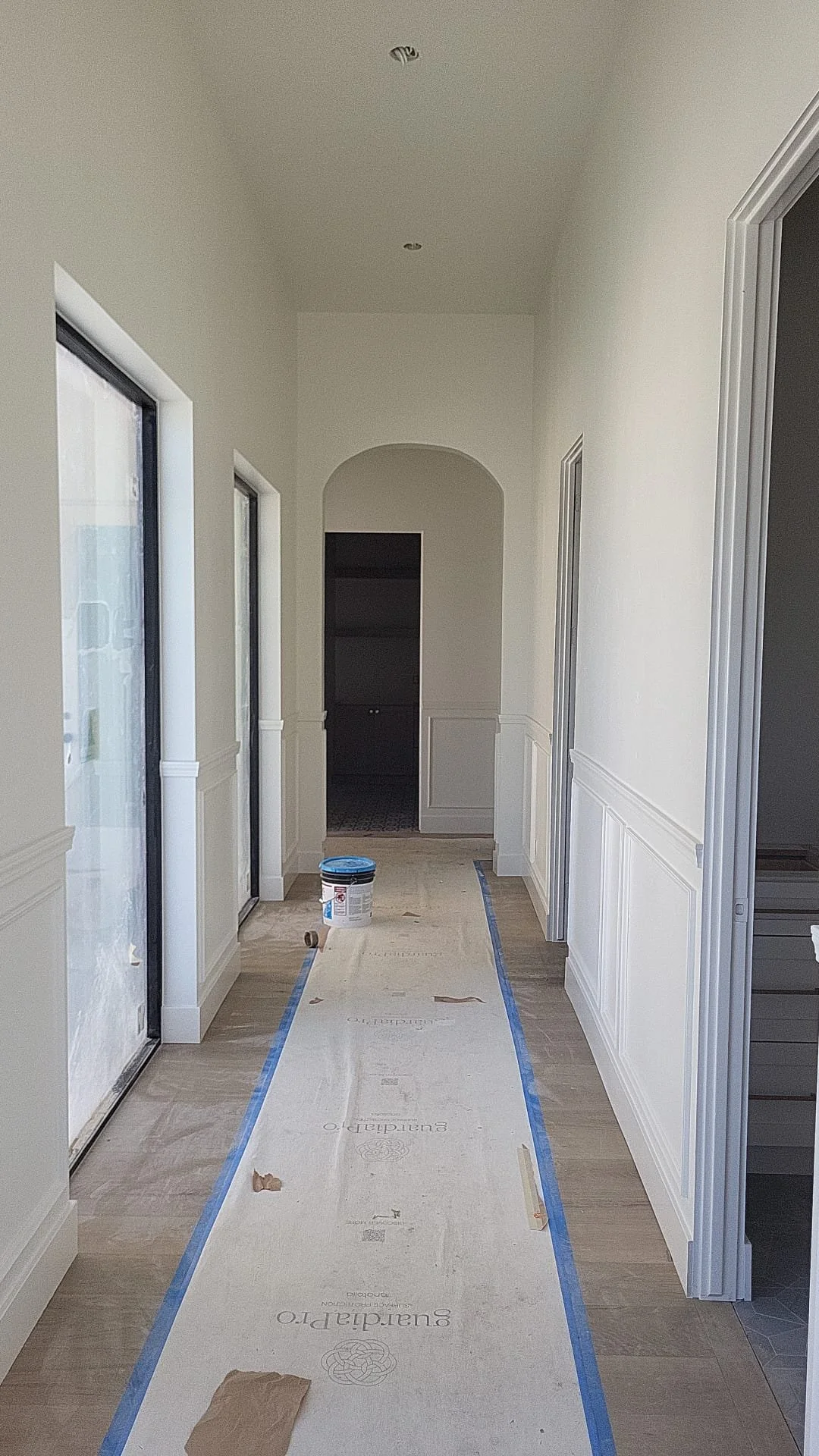A hallway under construction with white walls, large windows on the left, and doors on the right. The floor is covered with protective material and a bucket of paint is placed near the end of the hallway.