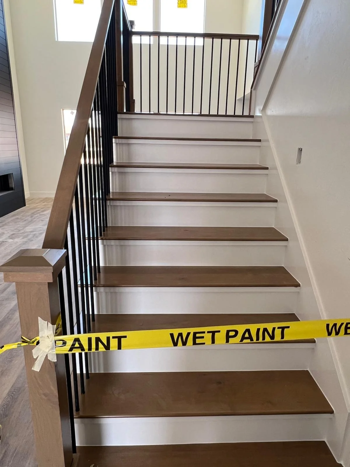 Interior view of a staircase with wooden steps and black metal railing, with yellow caution tape indicating wet paint across the bottom step.