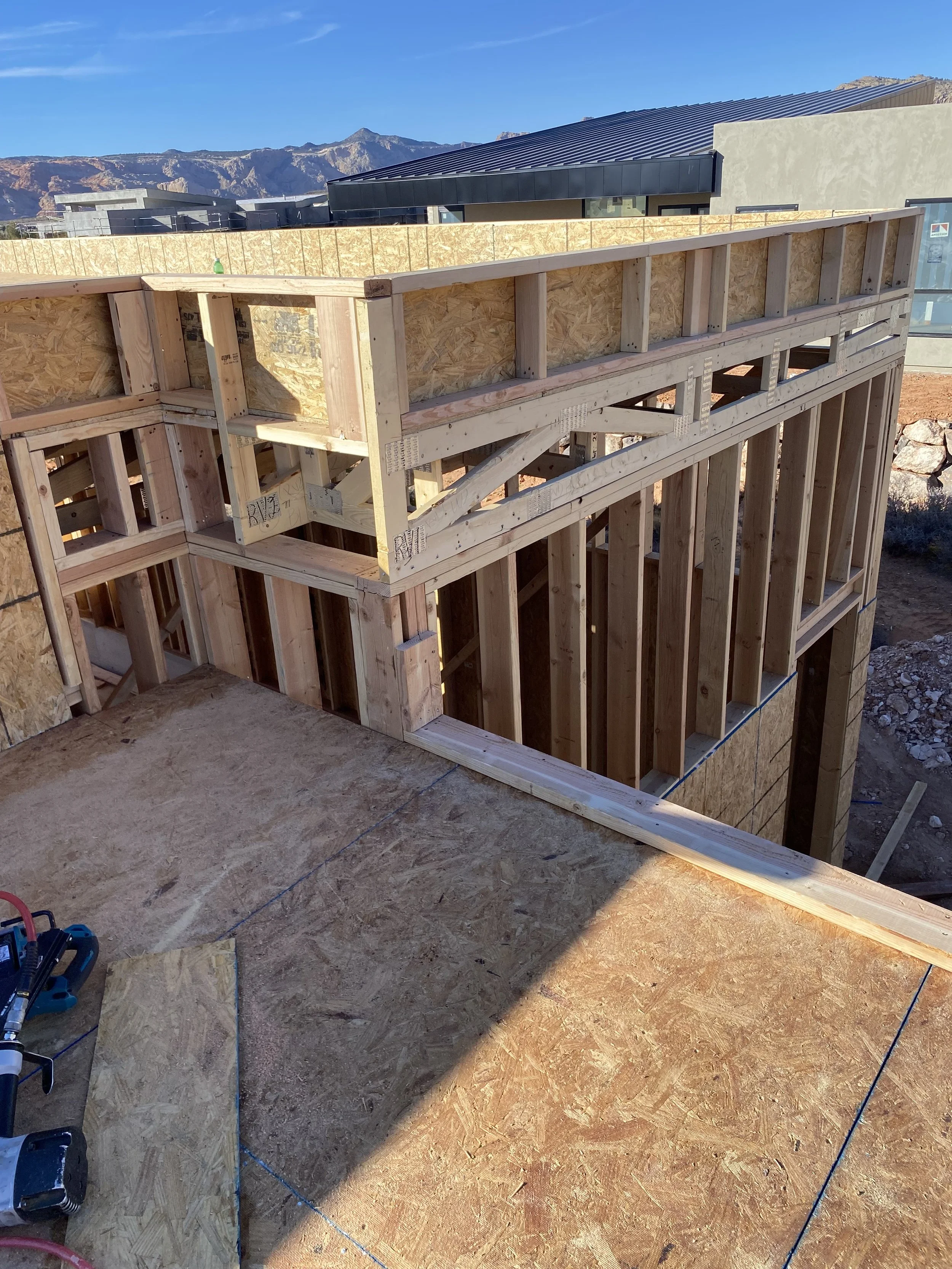 Construction site with a partially built wooden frame of a house or building, showing exposed beams and plywood sheathing, with mountains and a modern building in the background.