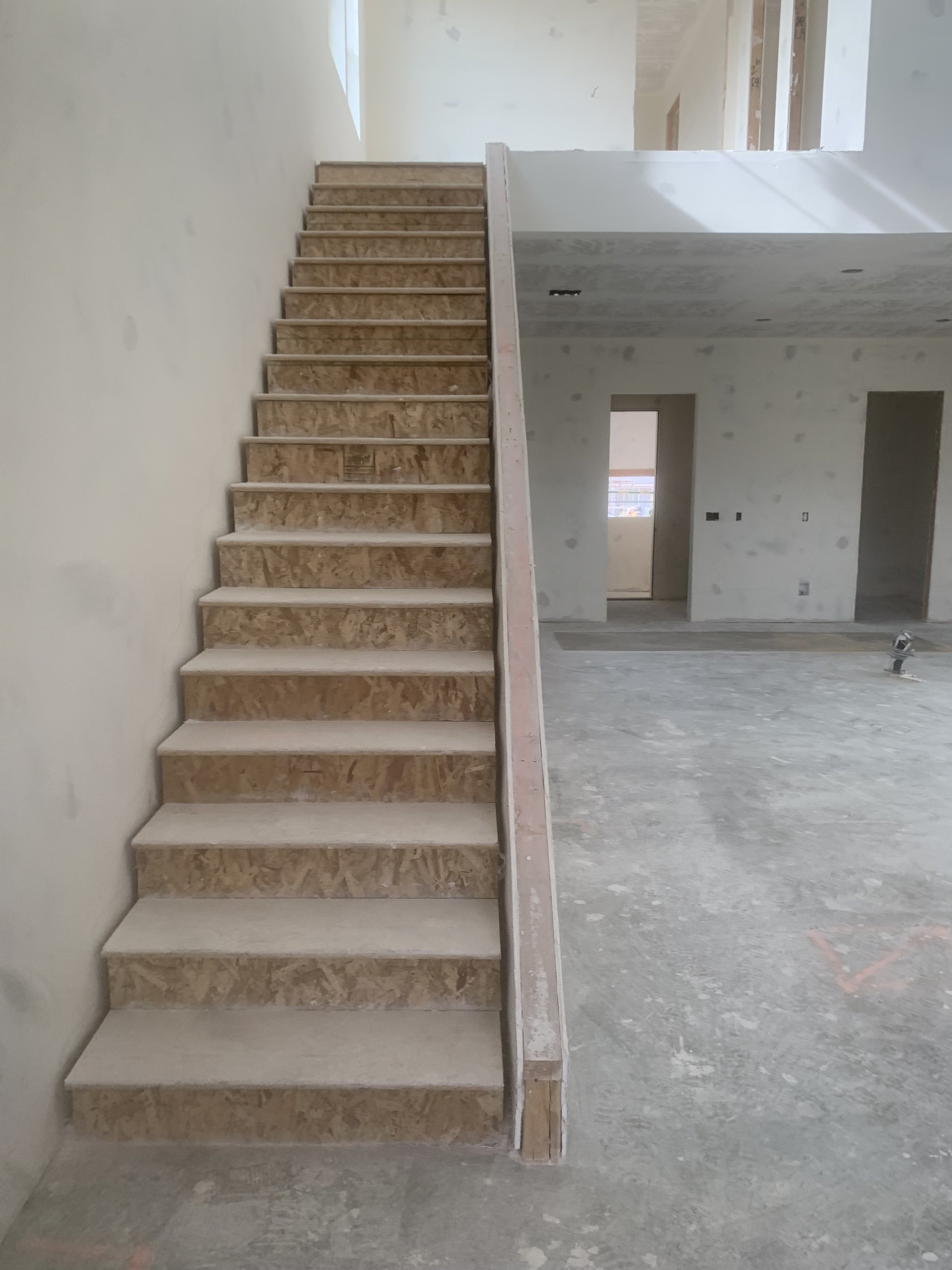 Unfinished interior of a house under construction with a wooden staircase leading to the second floor, surrounded by drywall walls and an open floor plan.