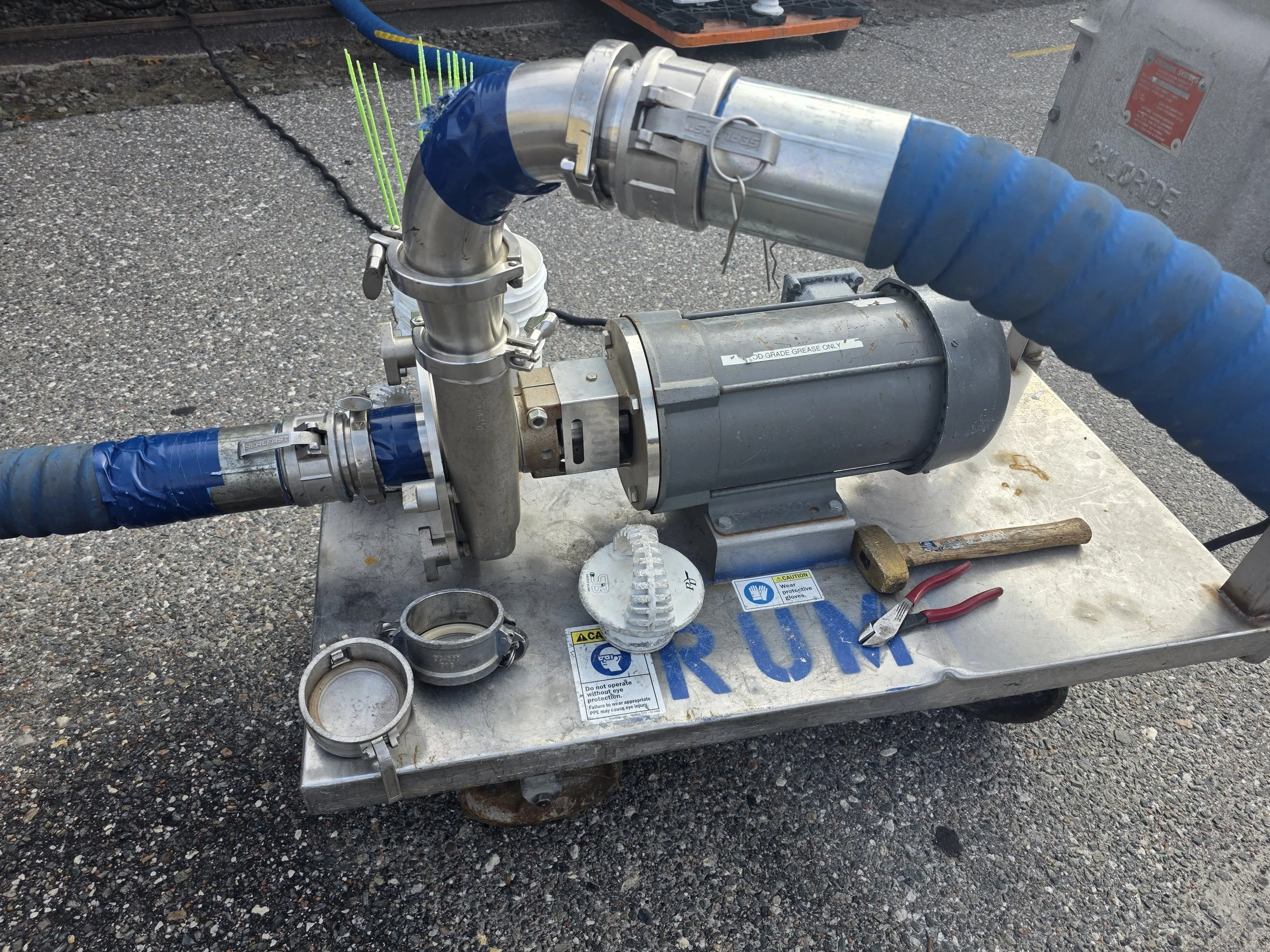 A metal pump with blue pipes connected, placed on a metal cart with the word 'RUK' written on it. Tools and fittings are nearby on the cart.