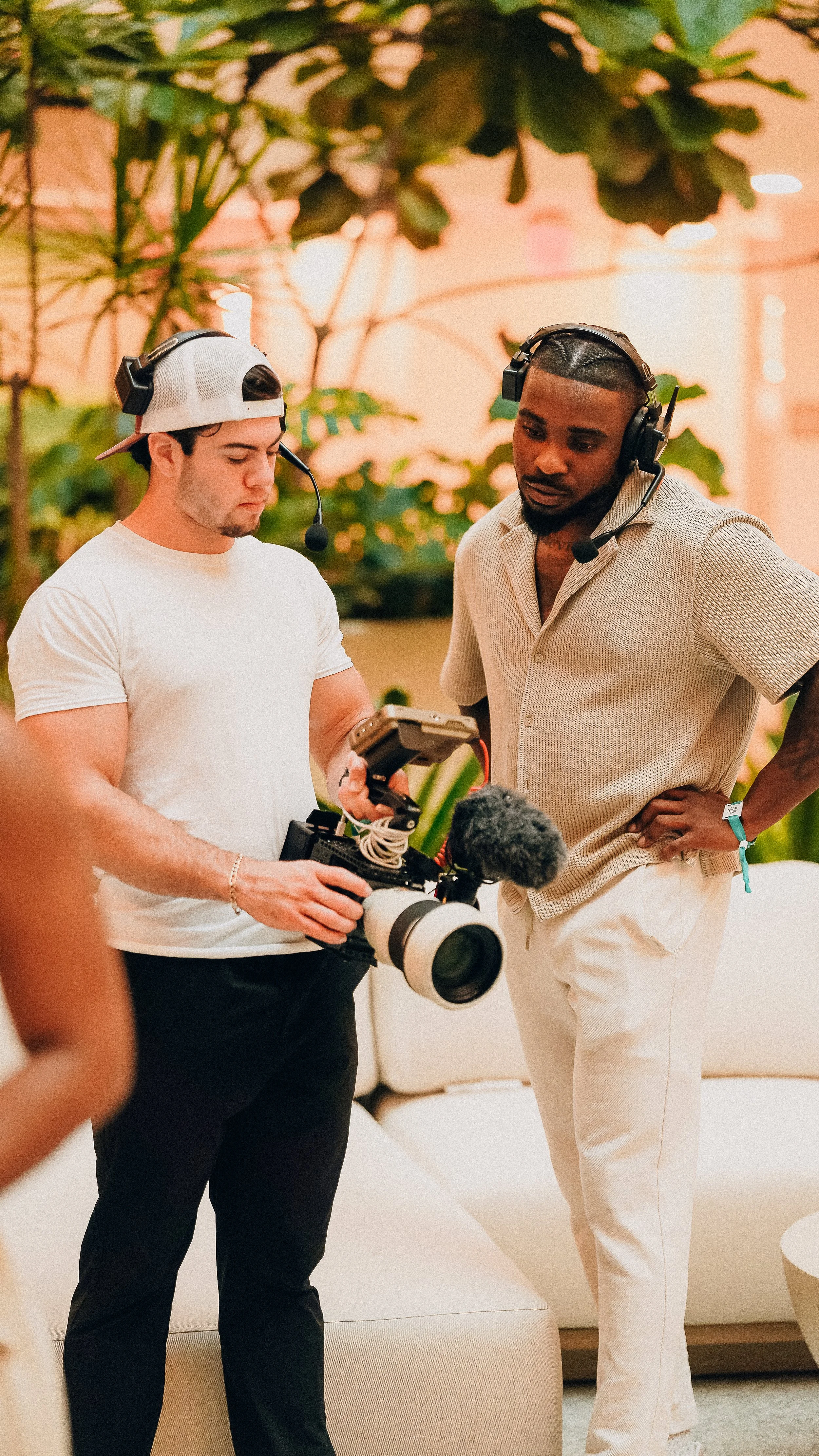 Two men wearing headsets review camera footage on a handheld monitor, standing in a room with tropical plants and peach-colored walls.