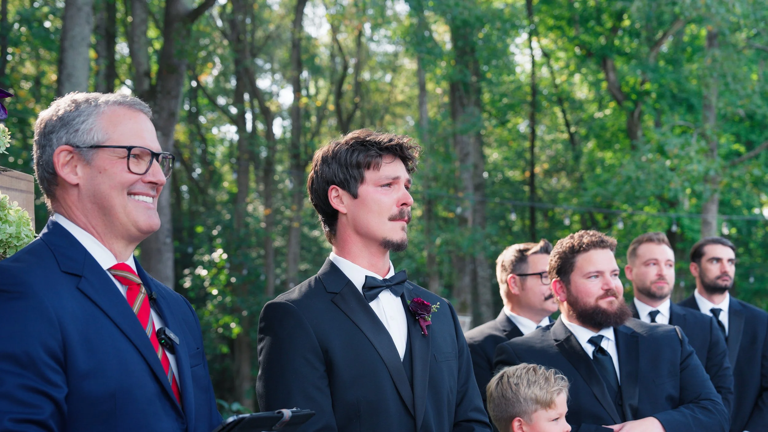 Group of men standing outdoors at a wedding ceremony, dressed in formal suits, with the groom in a tuxedo with a bow tie, and others in dark suits and ties, with trees in the background.