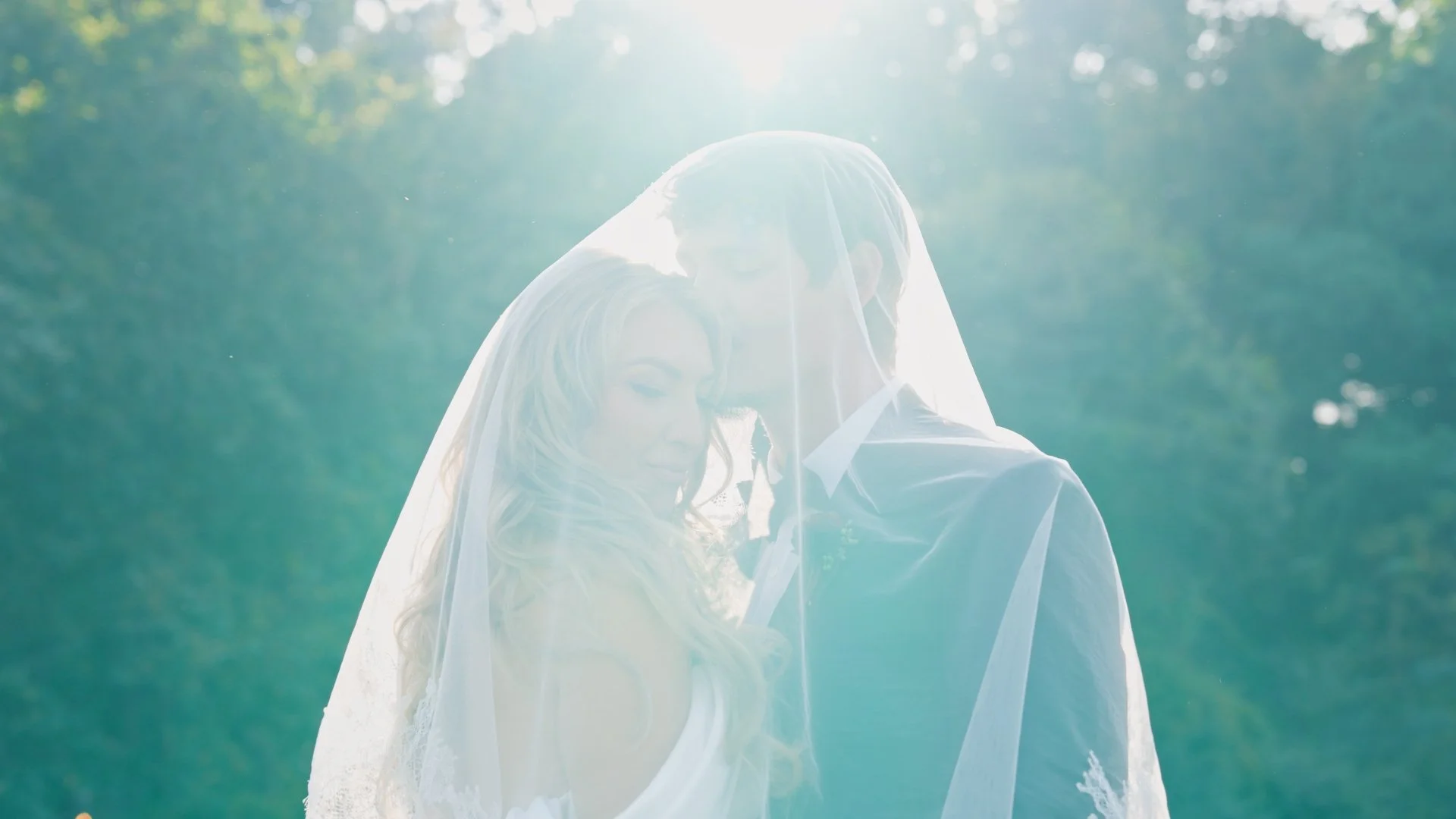 Bride and groom sharing a tender moment outdoors, with sunlight shining behind them and a lush green background.