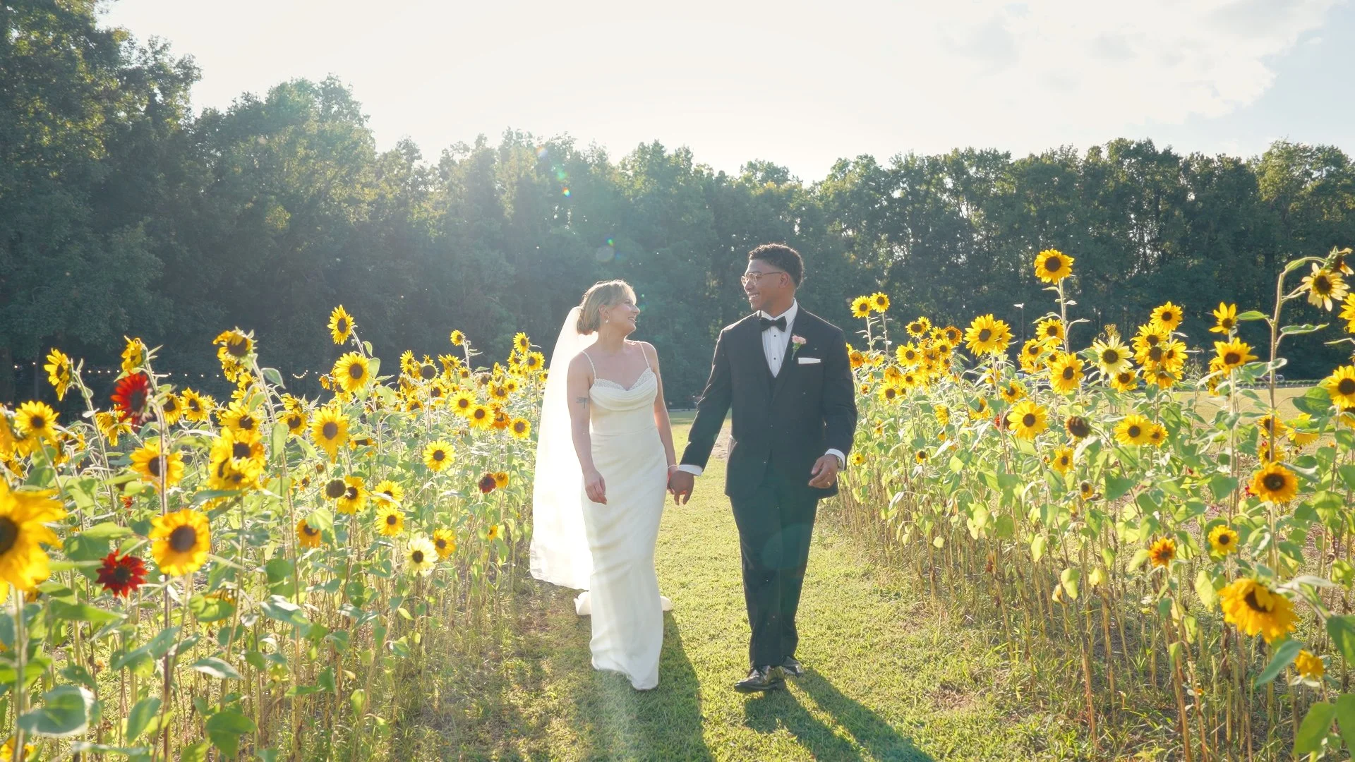 A wedding ceremony outdoors with groomsmen dressed in black suits and a man in a blue suit, red and gold striped tie, smiling, and a groom in a tuxedo with a purple boutonniere, standing among green trees.