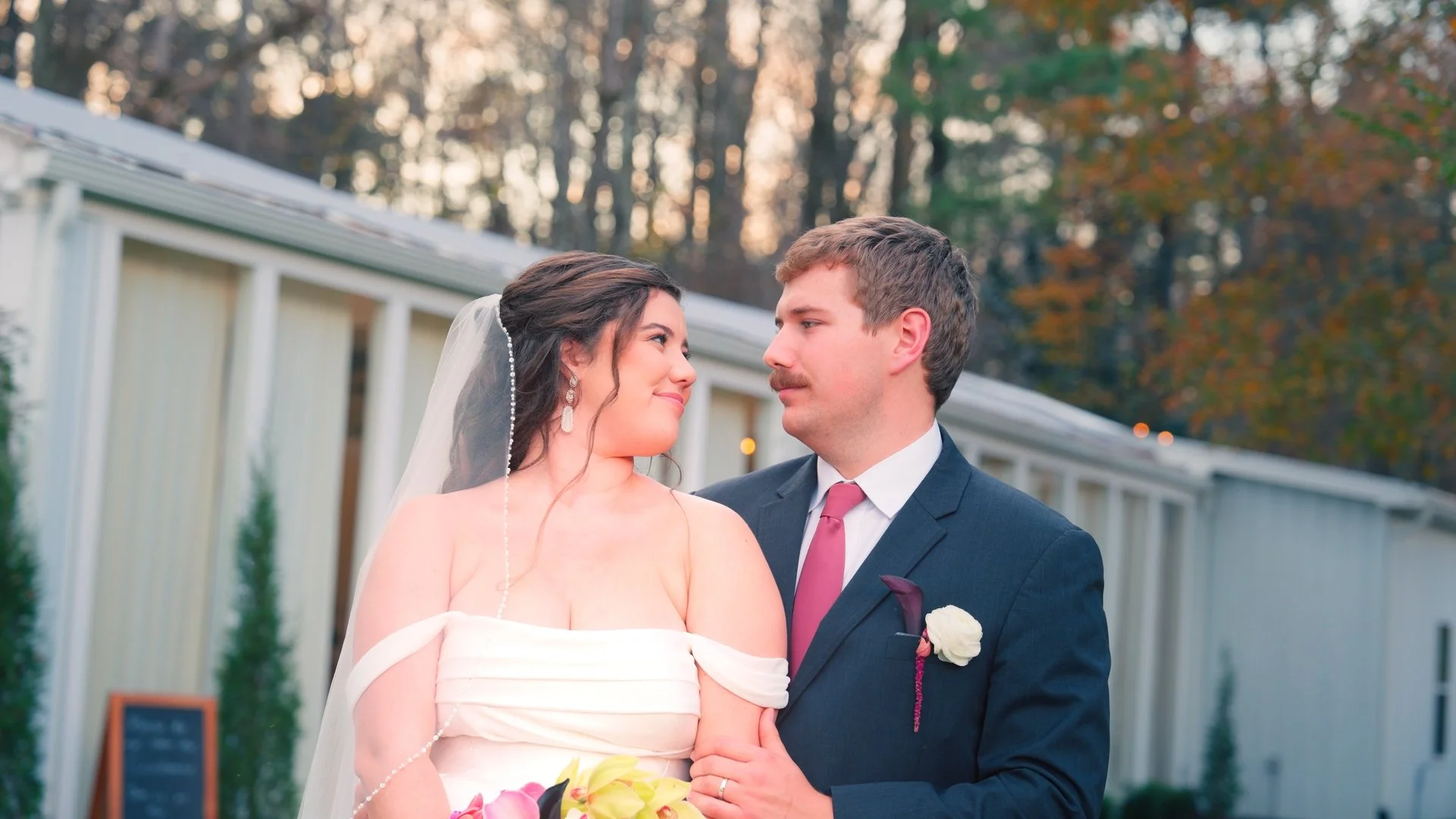 A couple in wedding attire sharing an intimate moment outdoors at sunset, with trees in the background.
