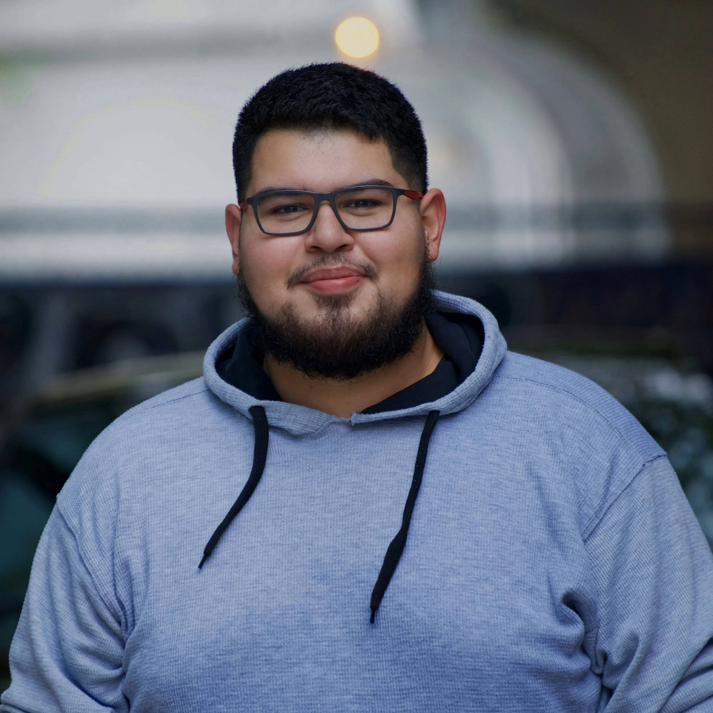 A young man with glasses, a beard, and a mustache wearing a grey hoodie, standing outdoors during sunset or sunrise.