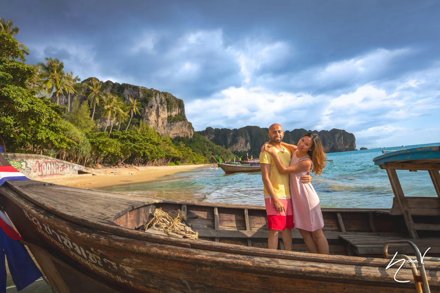 A couple beside a wooden boat with a tropical beach and cliffs in the background