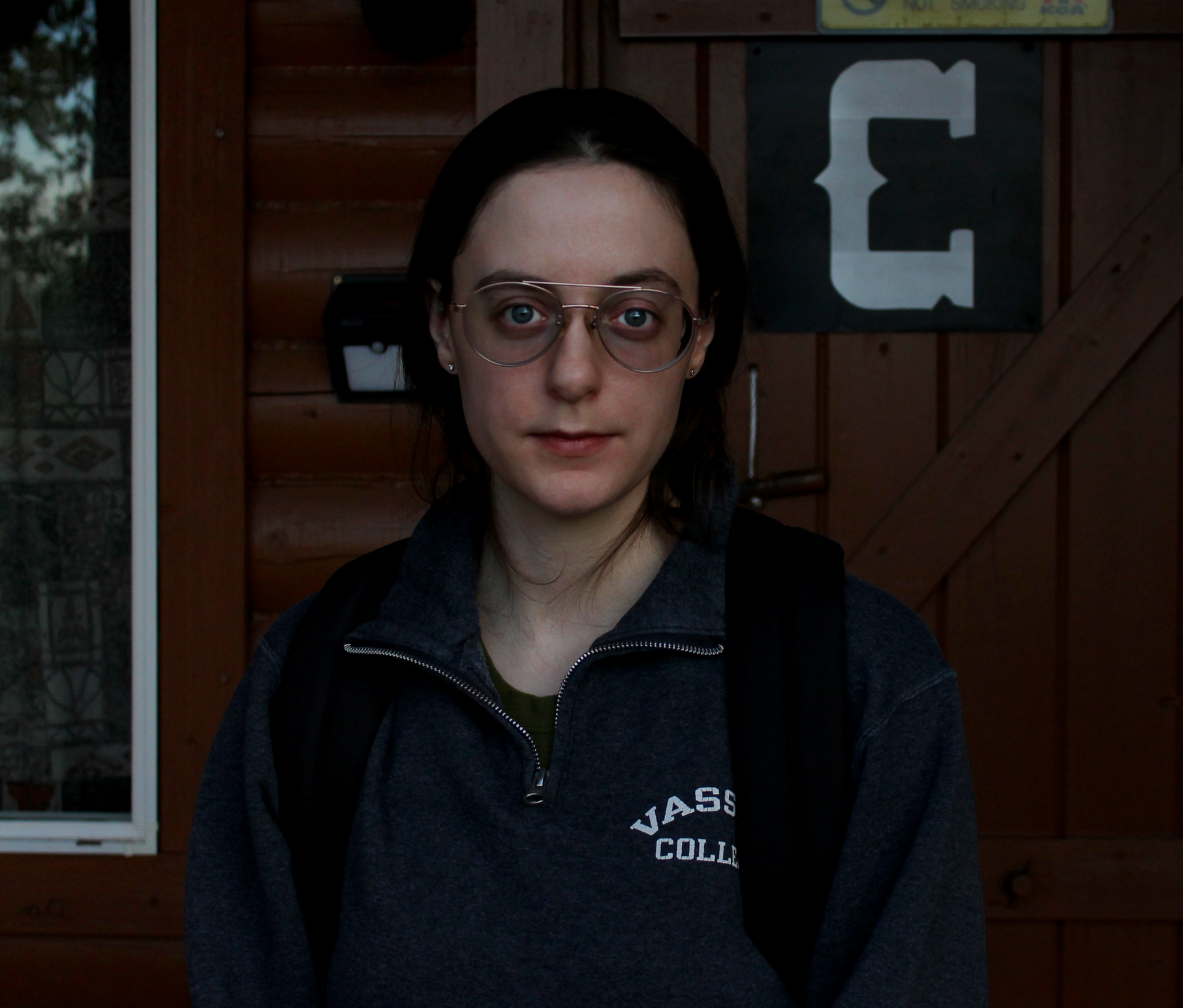 Candace M Osterhout, a young woman with dark hair, glasses, and a backpack standing in front of a wooden building, wearing a dark hoodie with 'VASSAR COLLEGE' written on it.