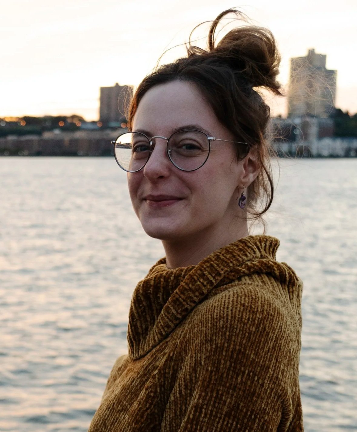 Candace M Osterhout, a woman with short hair and glasses standing by a body of water at sunset with city buildings in the background.