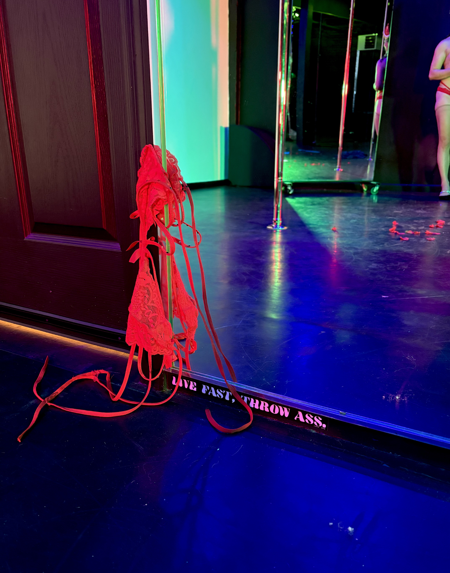 A lacy red bra hangs on the frame of a mirror above a sticker that reads "Live Fast, Throw Ass." Reflected in the mirror are a crhome pole and the left arm and leg of a standing light-skinned person.