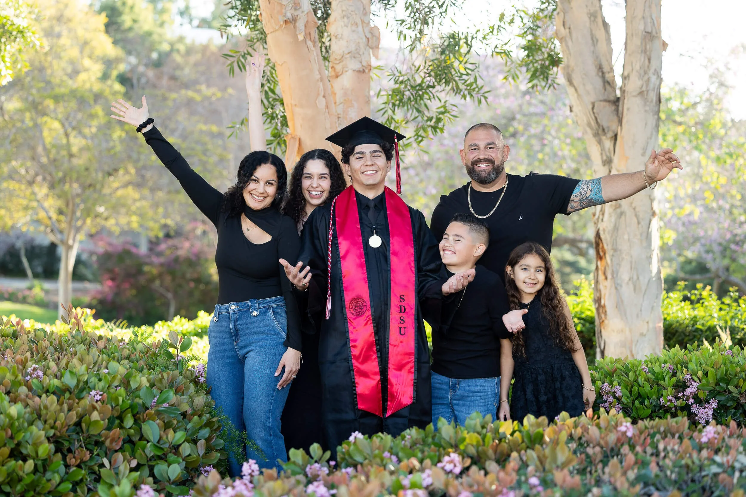 A family celebrating a graduation outdoors surrounded by greenery and trees, with two adults, two children, and a young man in a graduation cap and gown.