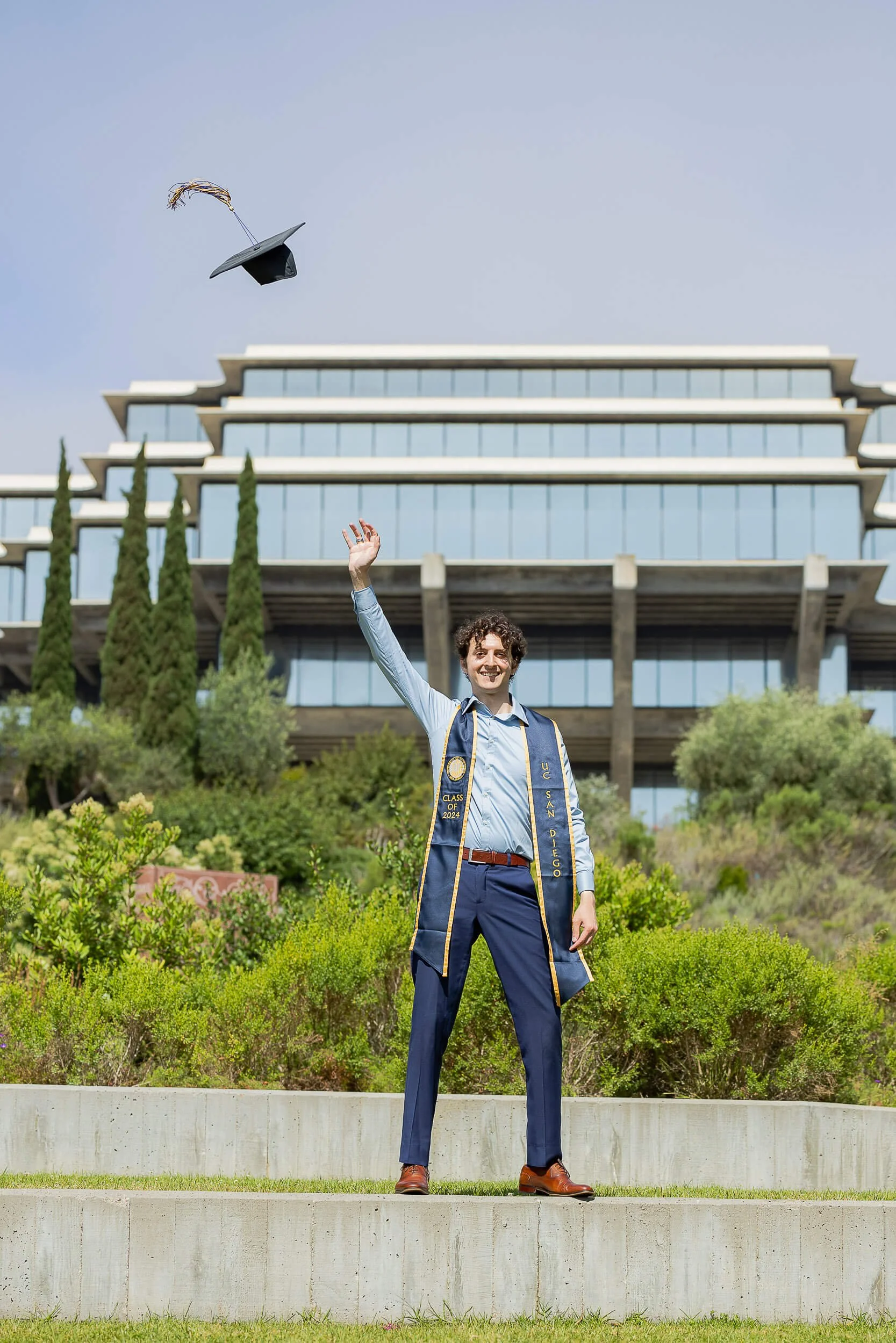 A male graduate wearing a blue gown and cap, standing outdoors on steps, throwing his cap in the air with a smile, in front of a modern building with greenery.