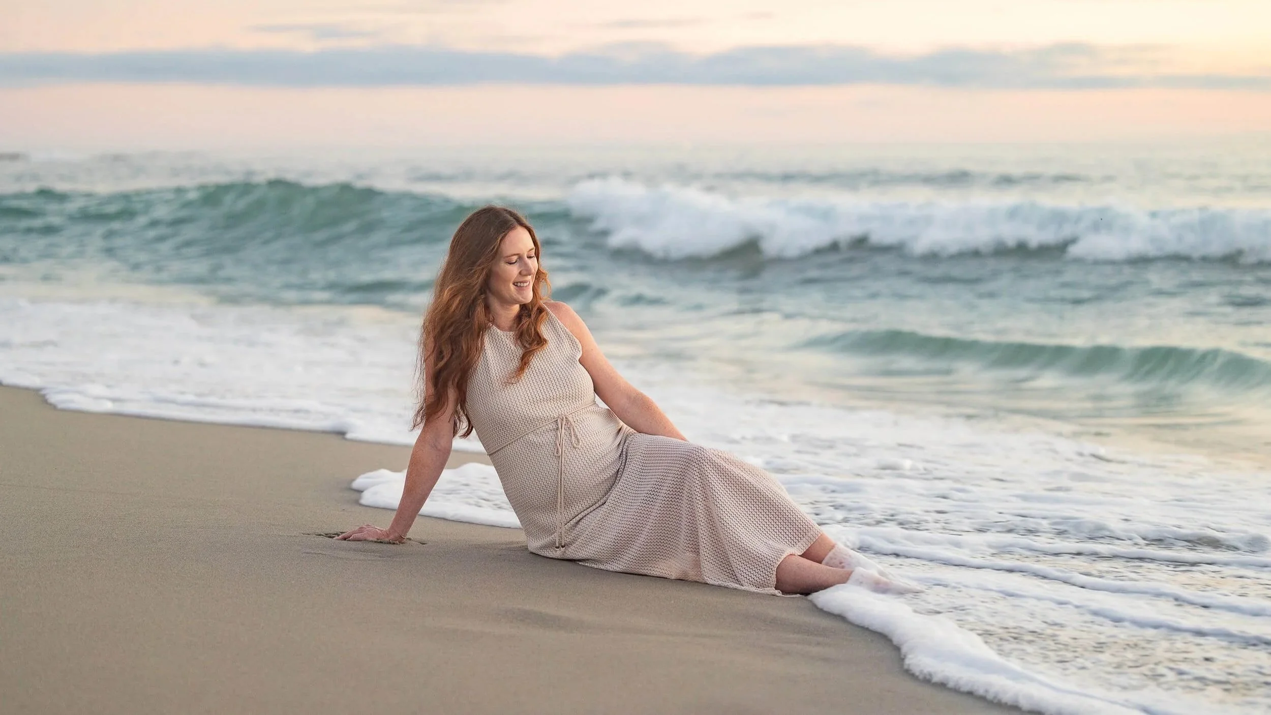 A woman sitting on the sandy beach near the shoreline, smiling and relaxing as gentle waves wash around her