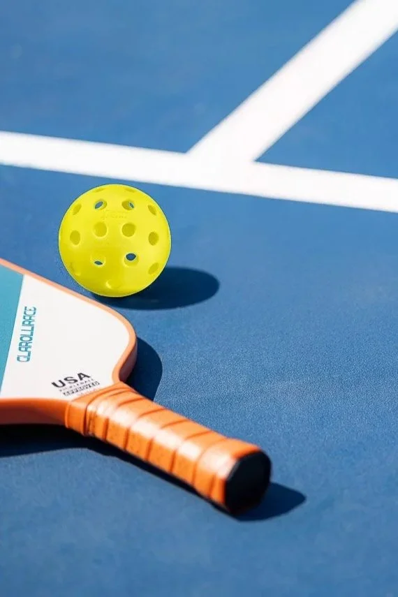 A yellow perforated tennis ball and an orange tennis racket with a black grip on a blue tennis court.