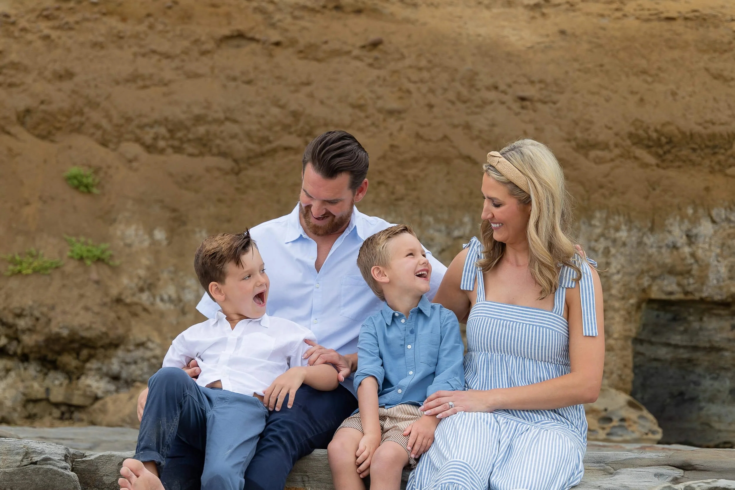 A happy family of four, a father, mother, and two young boys, sitting on rocks outdoors in front of a sandy hill, smiling and laughing together.