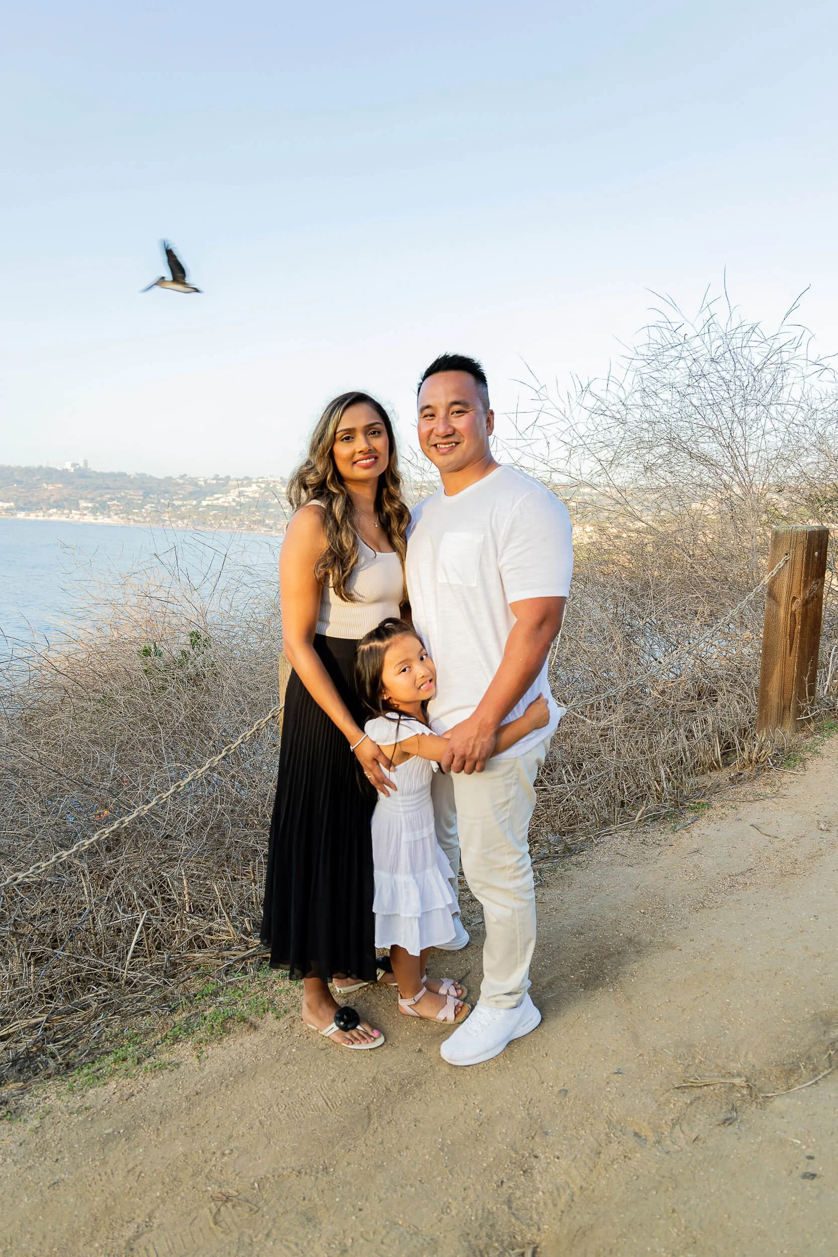 A family of four standing outdoors near a body of water, smiling at the camera. The background features dry bushes, a wooden fence, and a bird flying in the sky.
