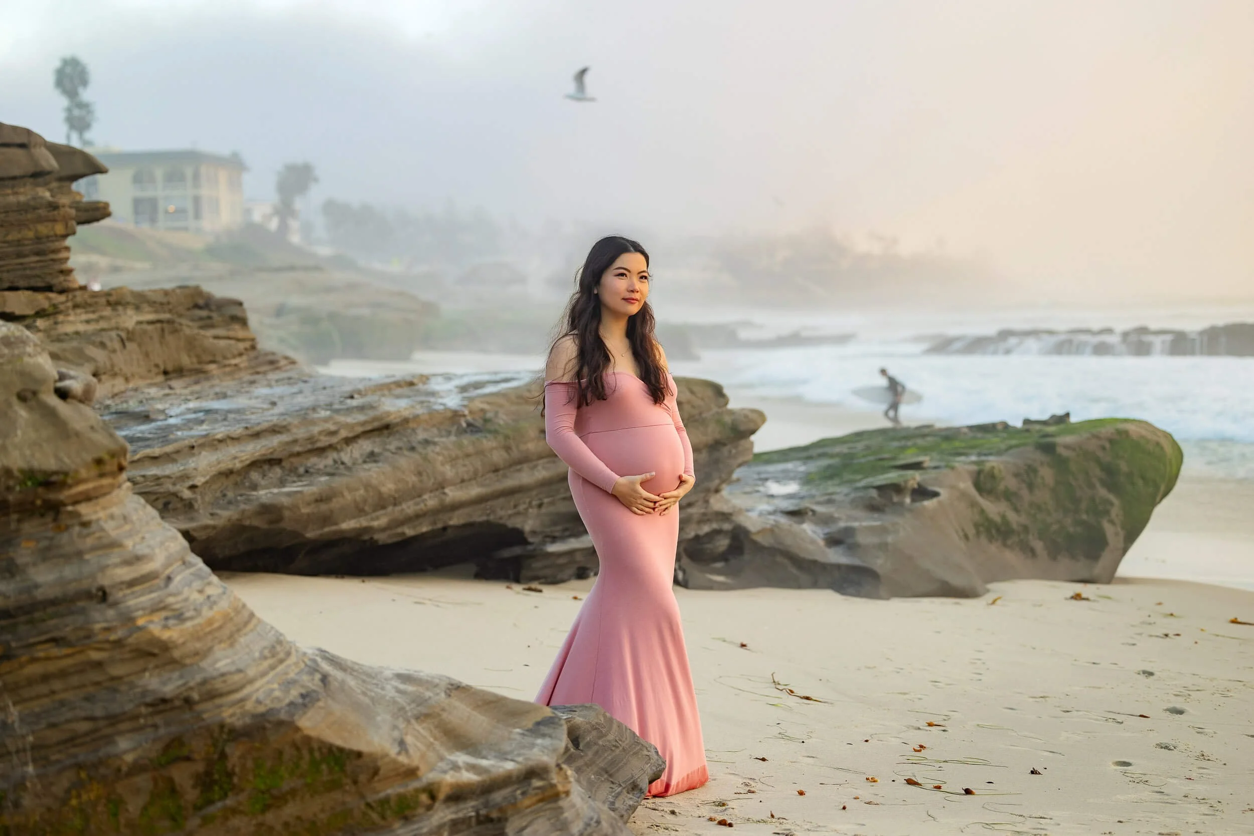 Pregnant woman in a pink dress standing on a sandy beach with rocks, ocean waves, and a surfer in the background, with a seagull flying overhead.