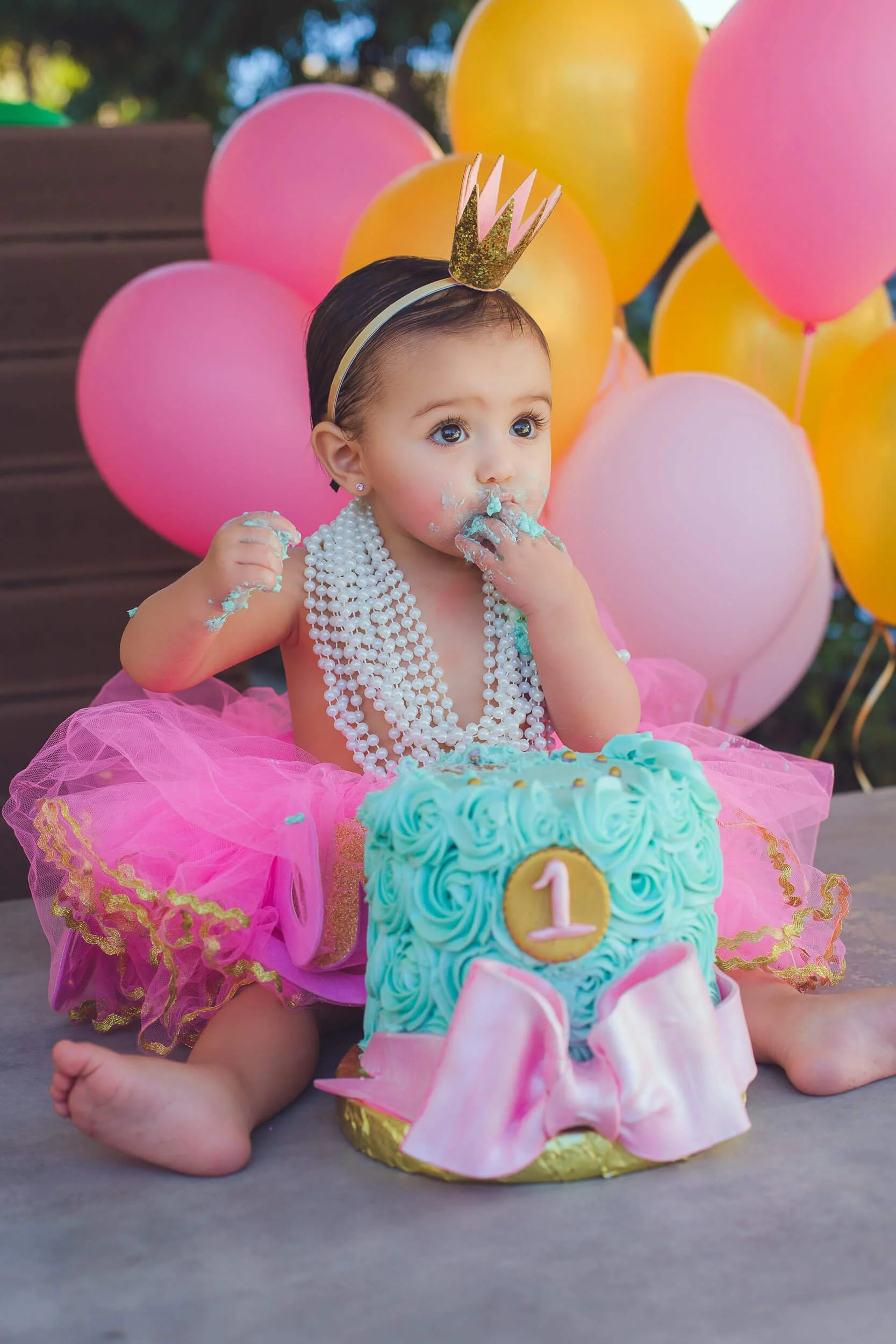 Baby girl wearing a pink tutu, pearl necklaces, and a gold crown headband, sitting in front of a blue birthday cake decorated with pink bow and a gold '1' sign, eating blue frosting, with multicolored balloons in the background.
