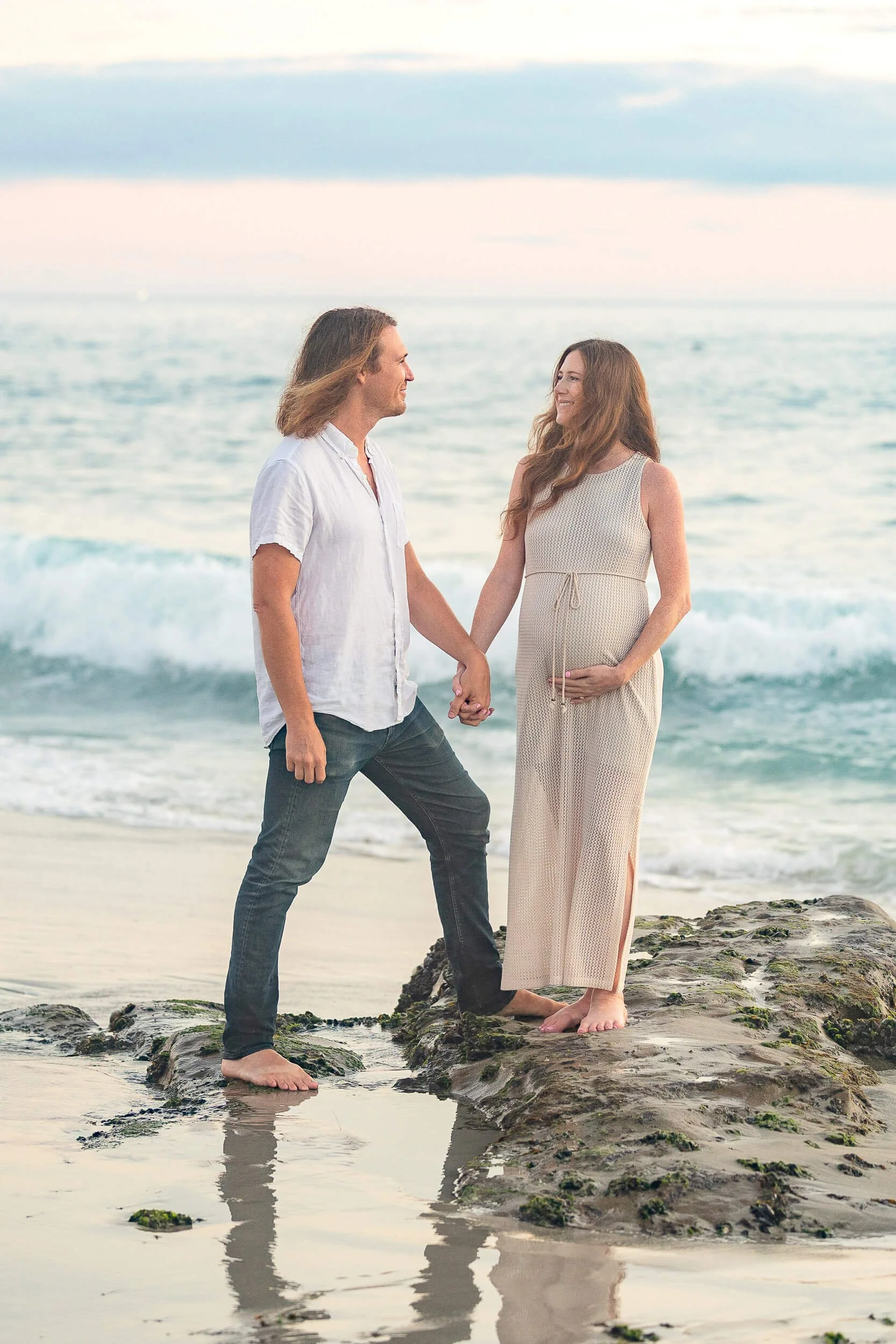 A pregnant woman and a man holding hands on the beach, standing on rocks with the ocean behind them, during sunset.