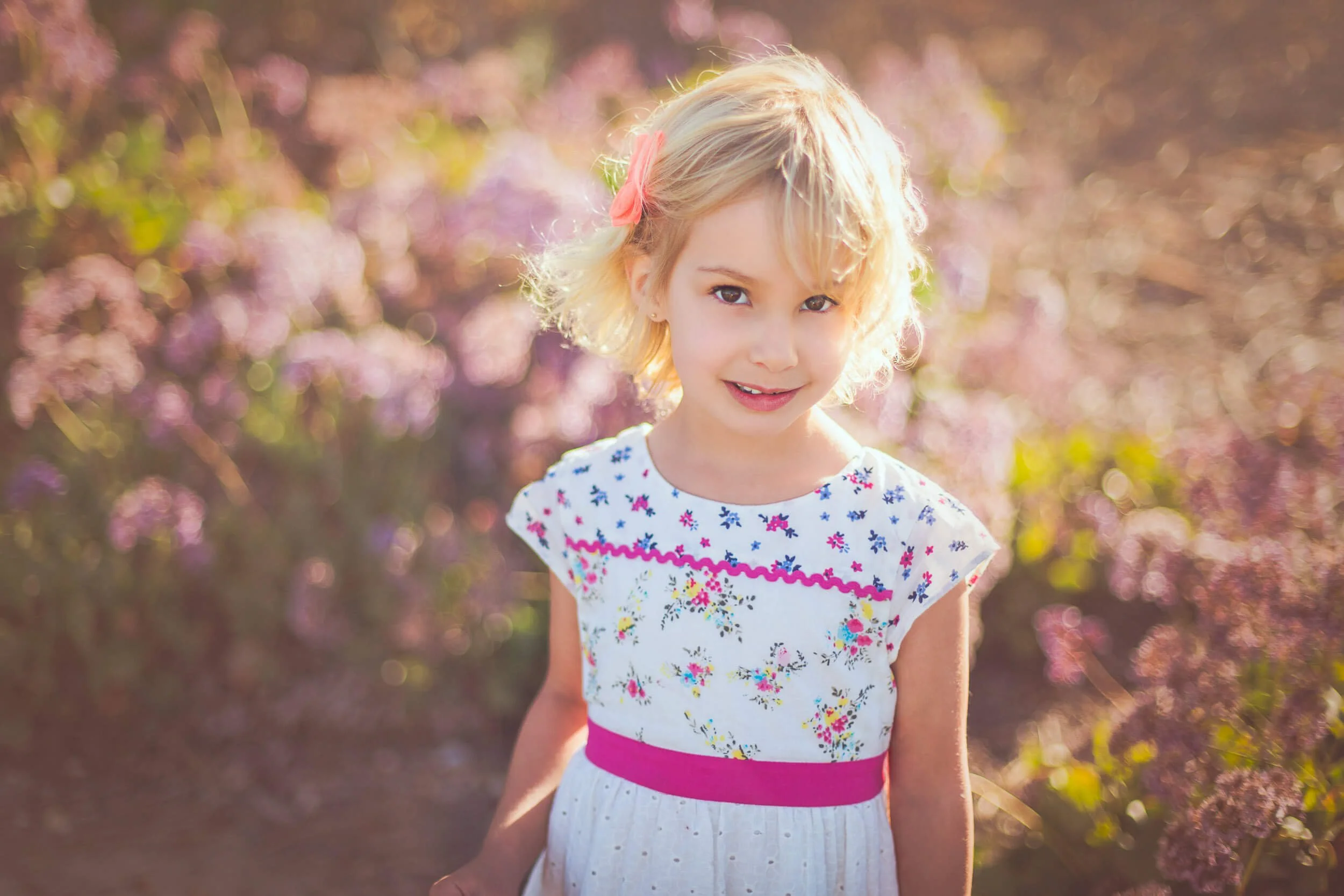 A young girl with blonde, curly hair and a pink bow in her hair, smiling outdoors with pink and purple flowers in the background.