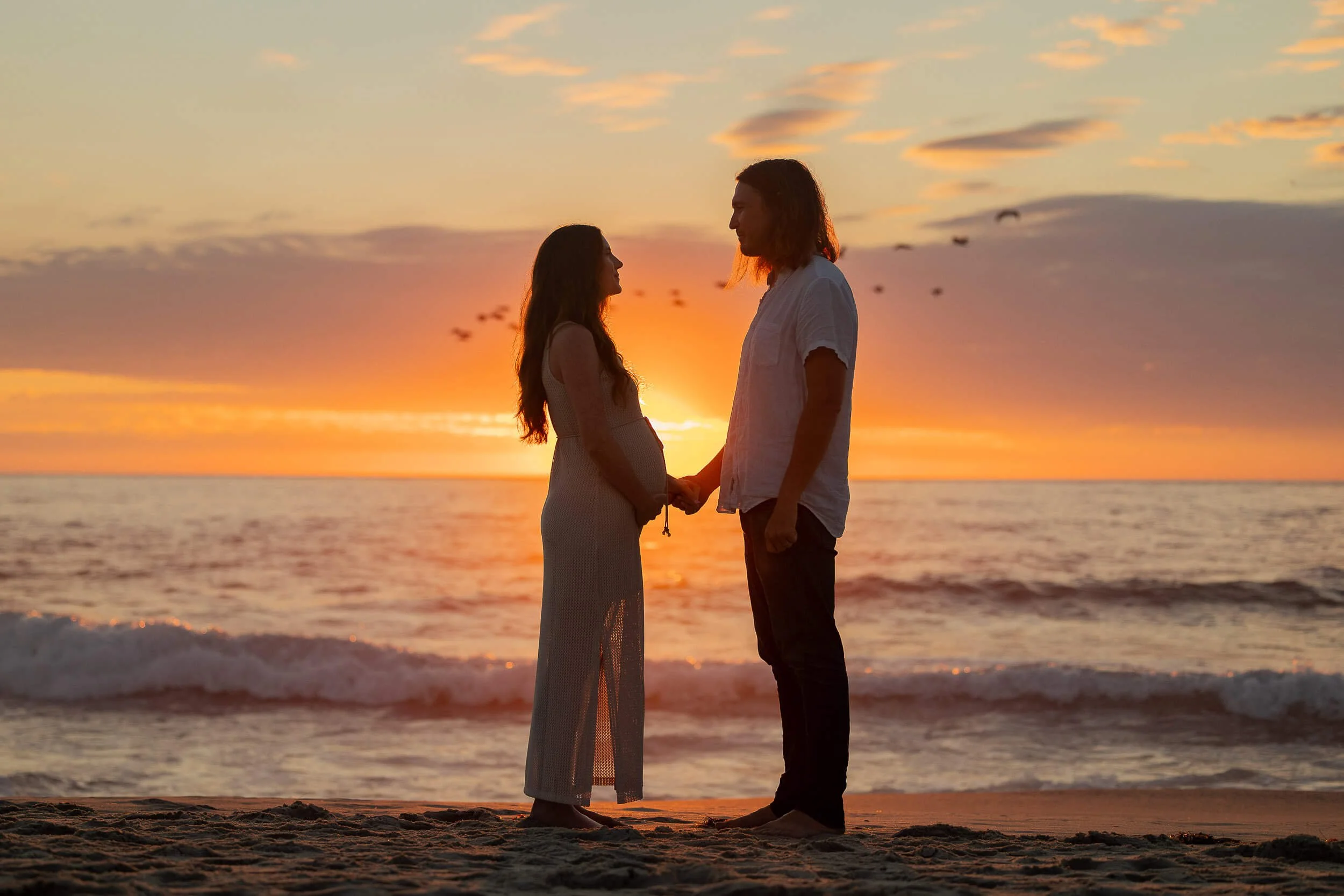 Silhouette of a pregnant woman and a man holding hands on the beach during sunset, with the ocean and colorful sky in the background.