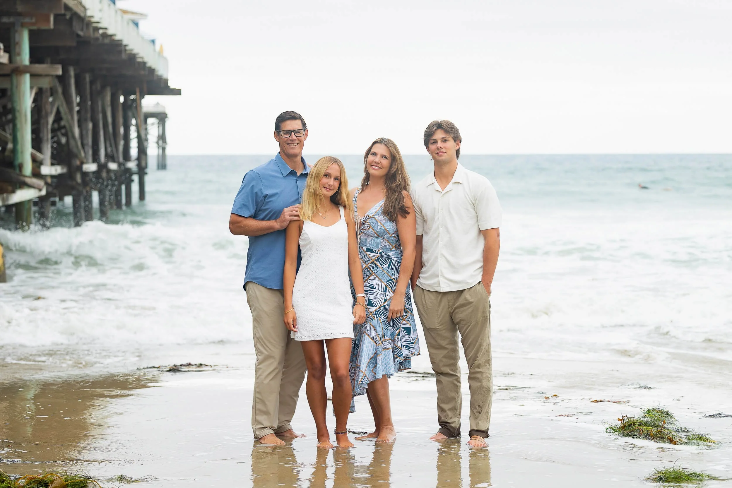 A family of four standing on the beach by the ocean, with a pier on the left. The family members include a man, a woman, a teenage girl, and a teenage boy, all smiling and dressed in casual, beach-appropriate clothing.