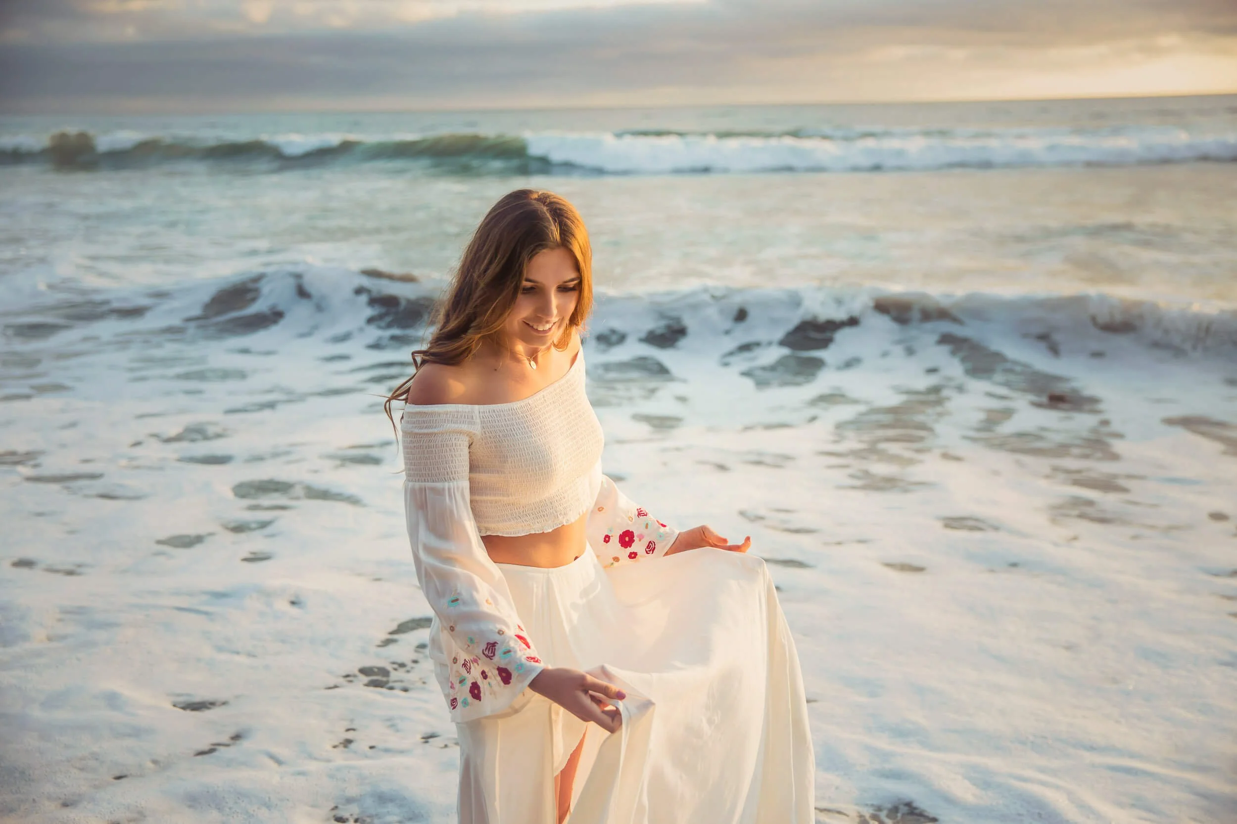 A young woman stands at the beach with waves in the background, smiling and holding up her flowing white skirt.