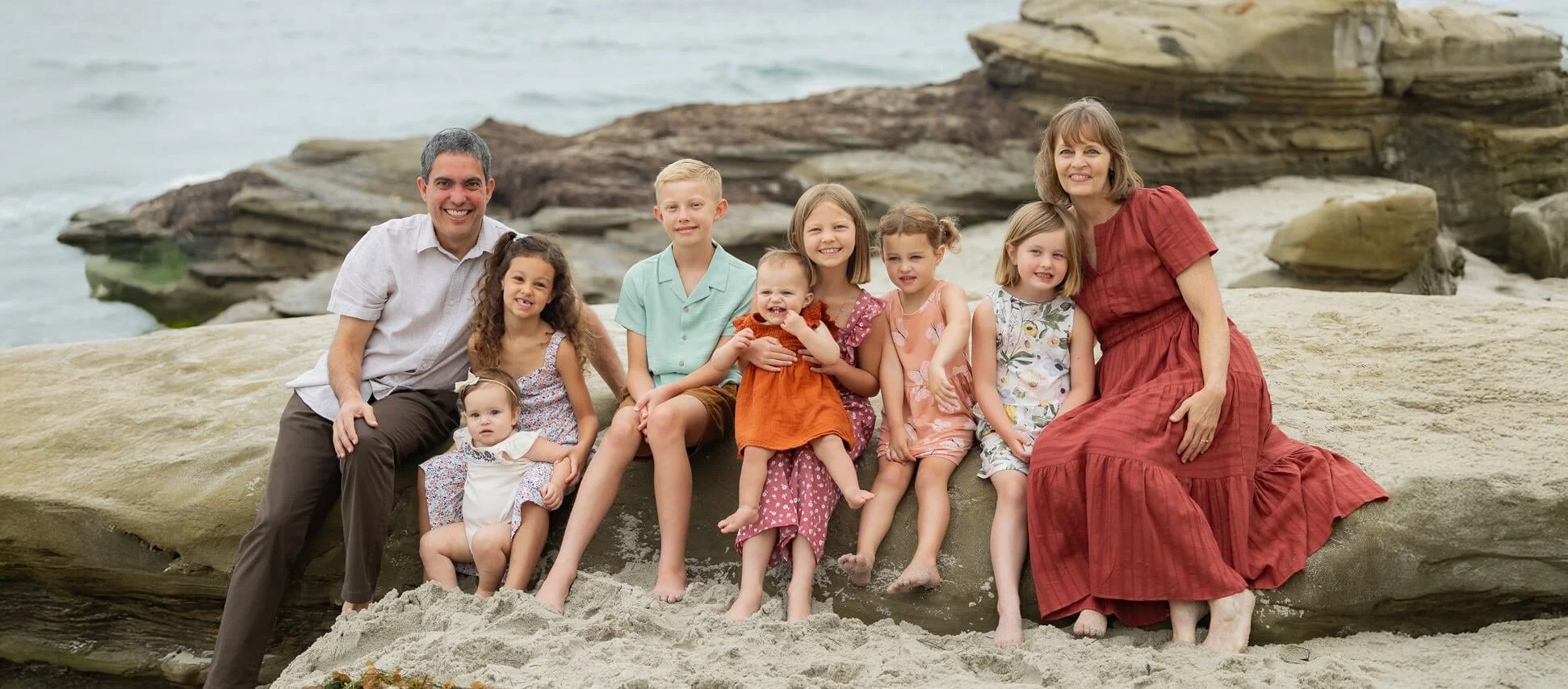 Family of ten sitting and posing on rocks by the beach with ocean in the background, smiling at the camera.