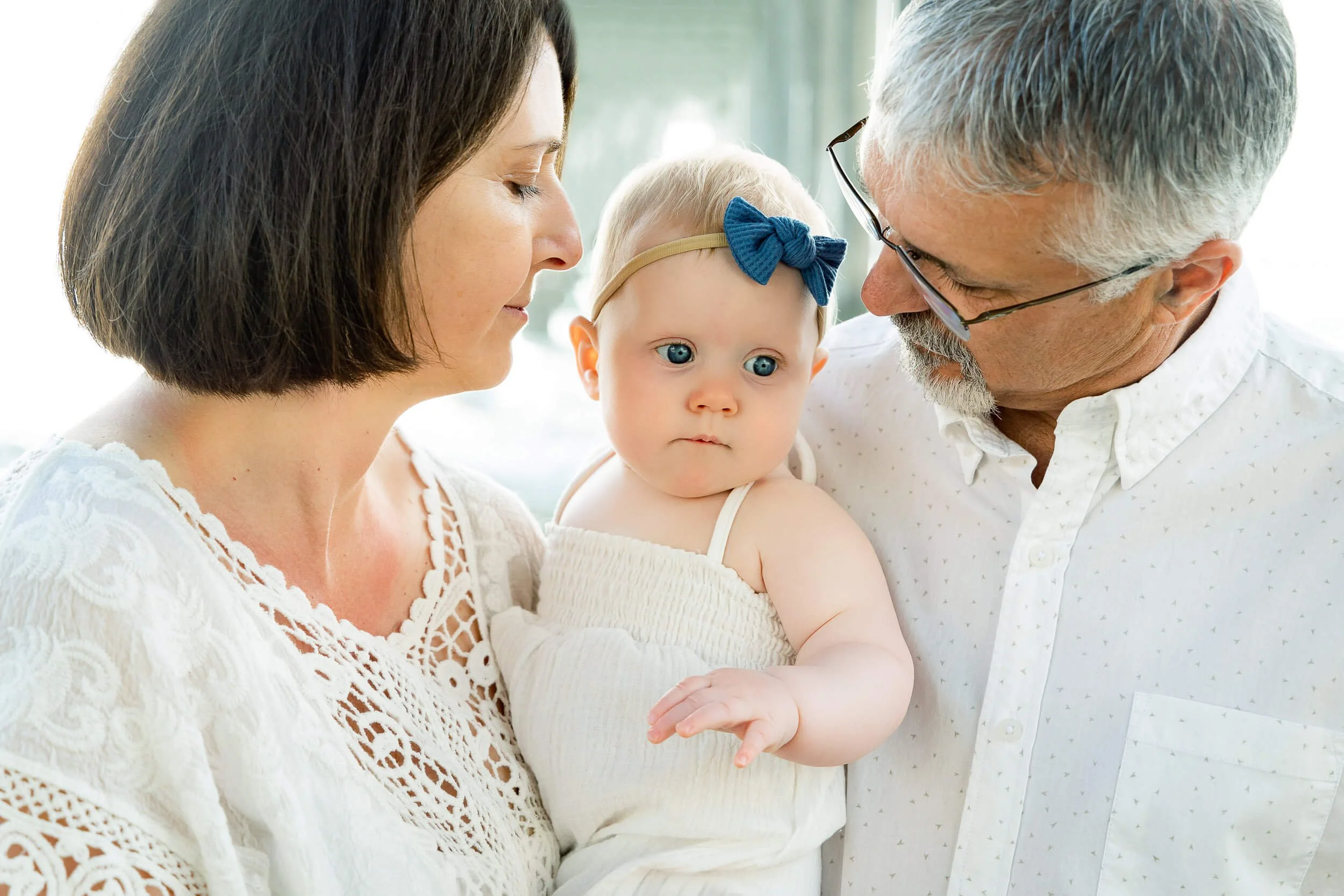 A woman and an older man holding a baby girl, all with light skin, indoors. The woman has short brown hair and is wearing a white lace top, the man has grey hair and glasses, and the baby girl has blue eyes, light blonde hair with a navy blue bow hea