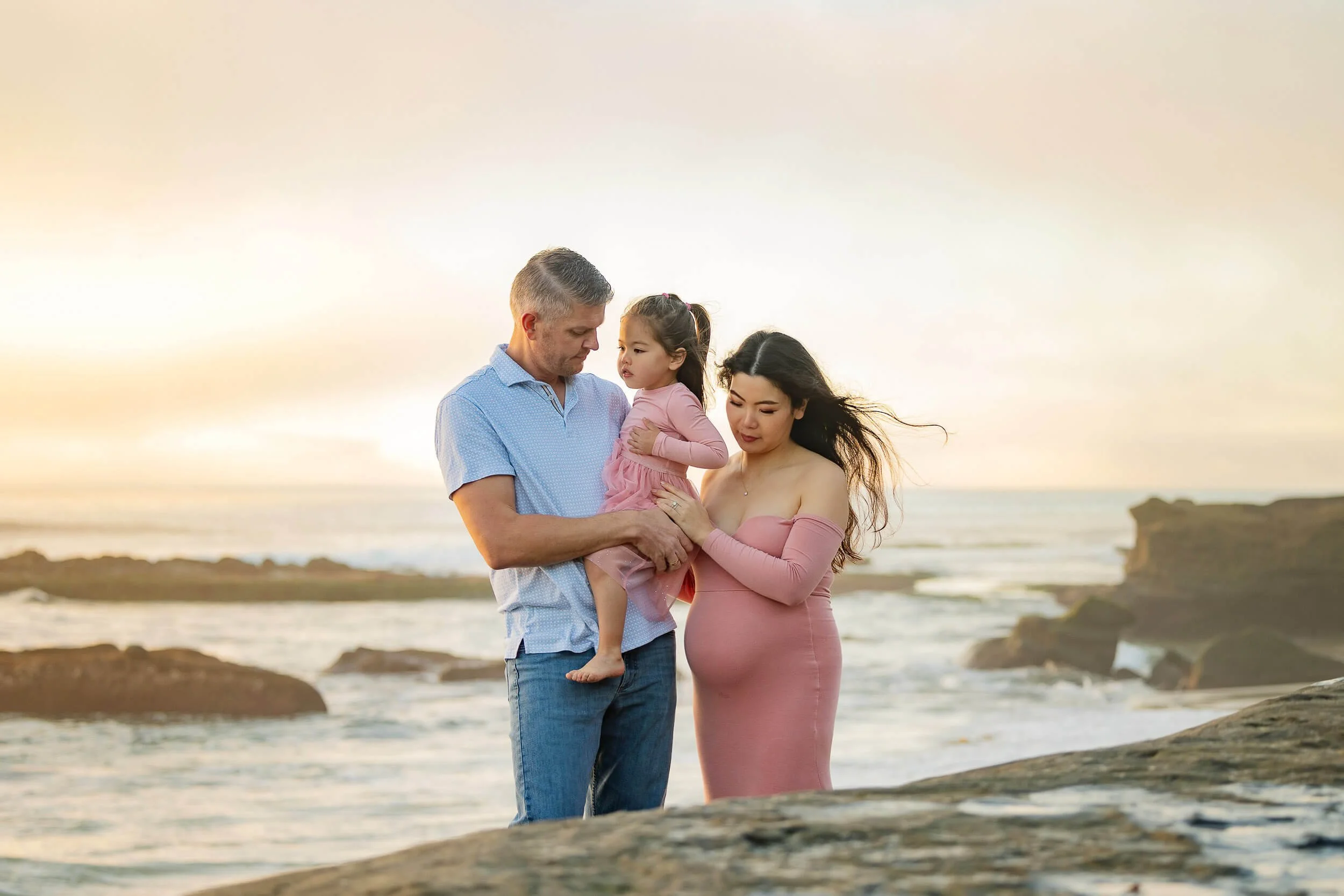 A family of three on the beach during a sunset, with the father holding his young daughter while the mother looks on, all standing on rocks near the ocean.