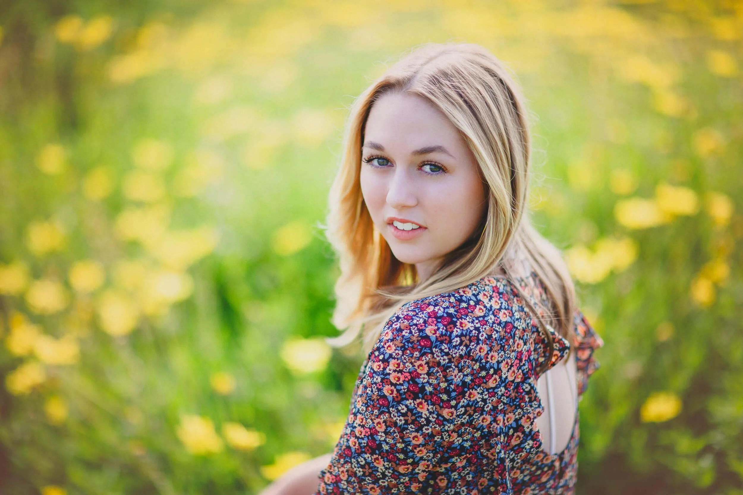 A young woman with blonde hair and blue eyes looking over her shoulder in a floral dress outdoors with blurred yellow and green foliage in the background.