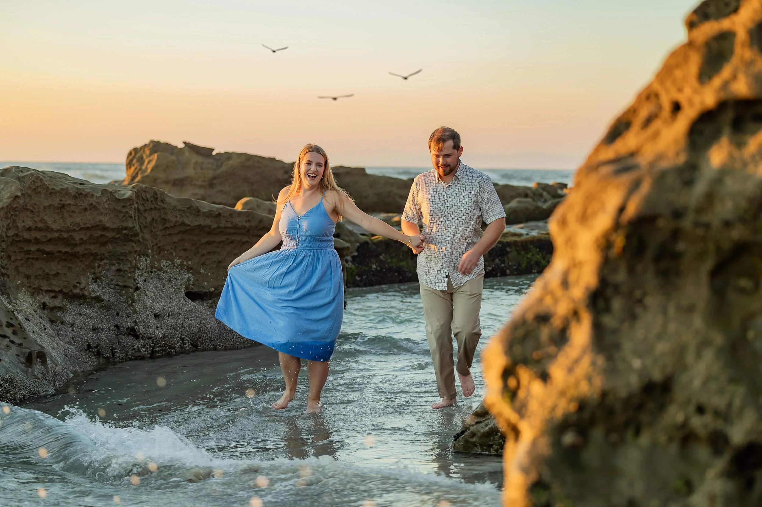 A smiling woman in a blue dress and a man in a patterned shirt and beige pants holding hands while walking barefoot in shallow ocean water among rocks during sunset
