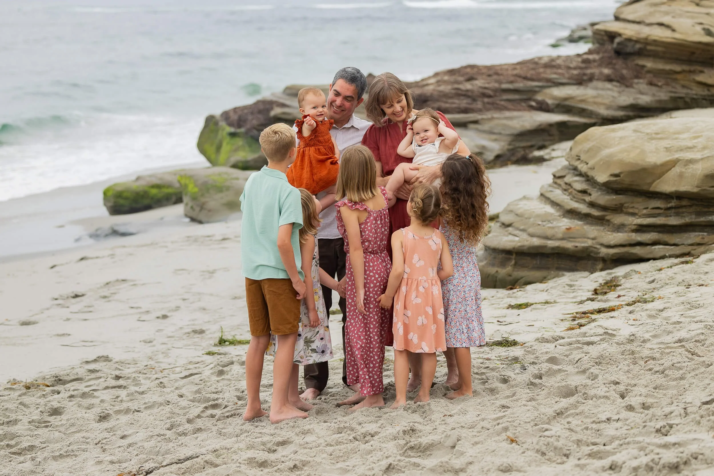 A family of seven, including two babies, on a beach with rocks and ocean waves in the background.