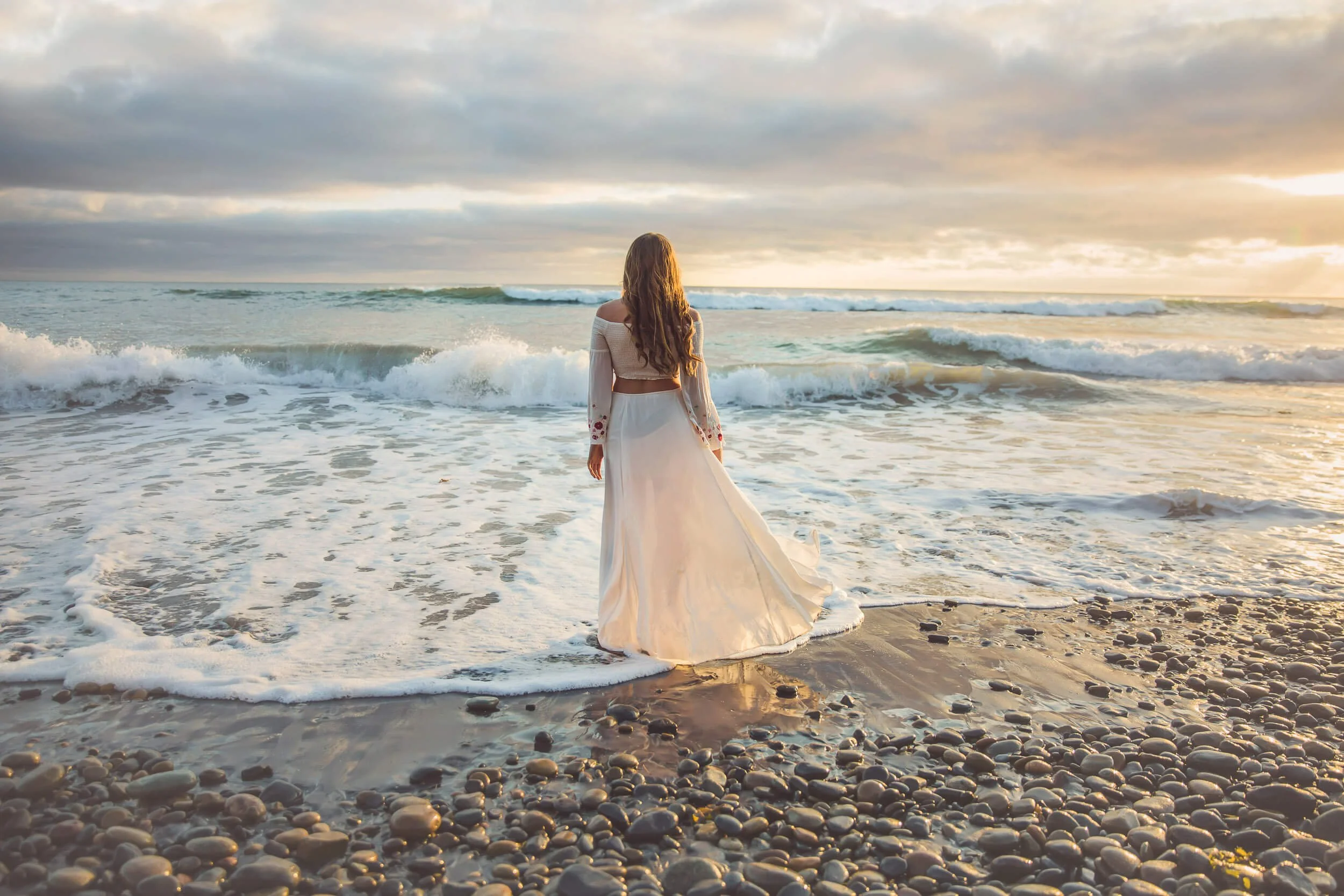 A woman in a white dress standing on a pebble beach looking at the ocean at sunset.