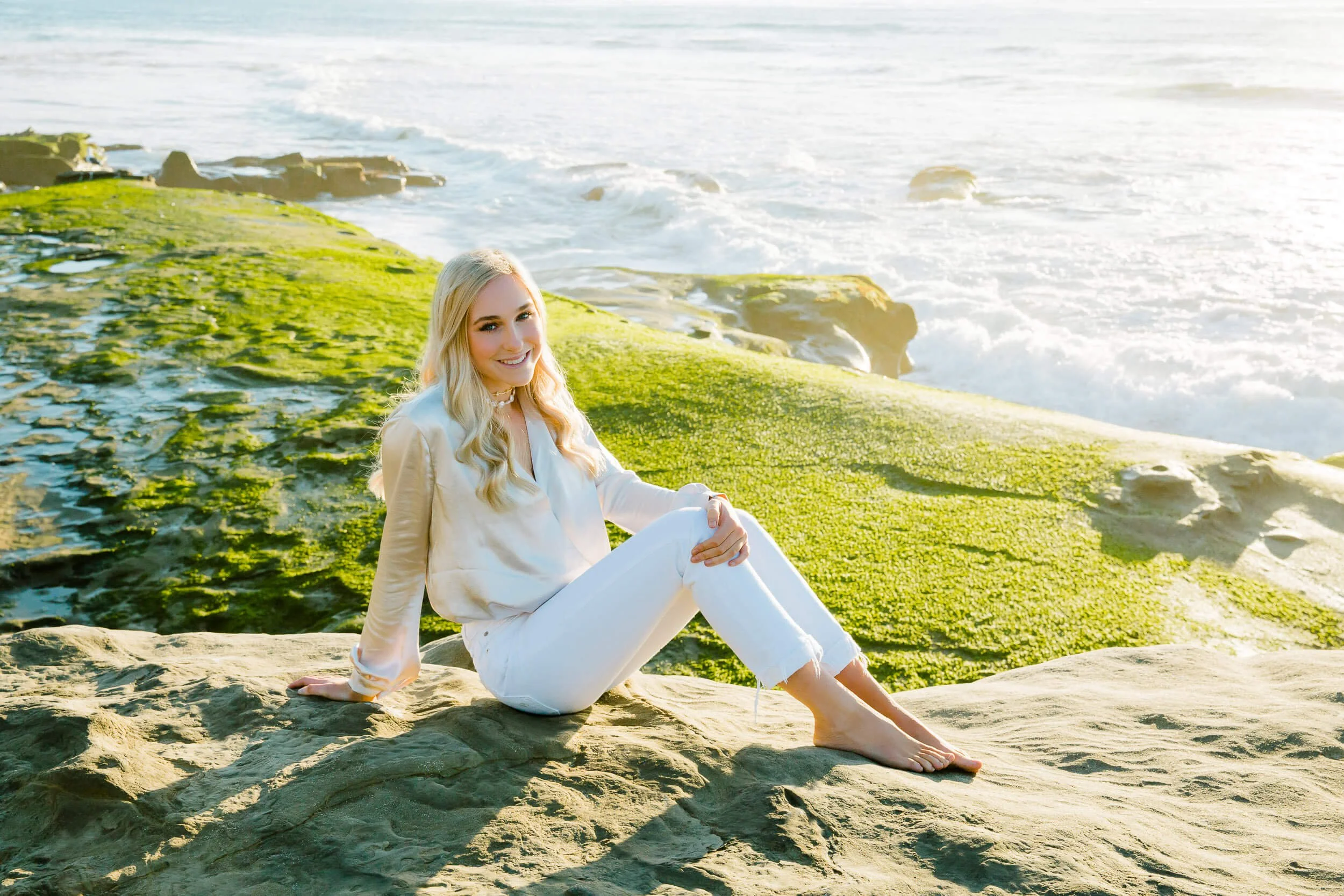 A young woman with blonde hair wearing a white outfit sitting on a rock by the green moss-covered shoreline with ocean waves in the background.
