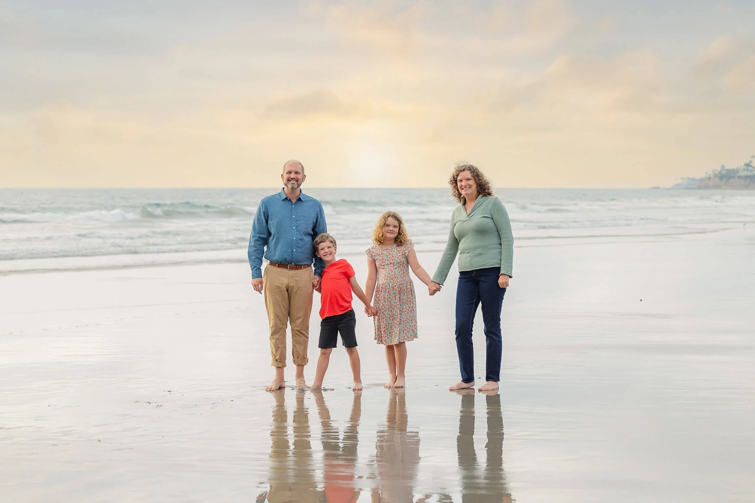 Family of four standing on the beach holding hands, with the ocean and a sunset in the background.