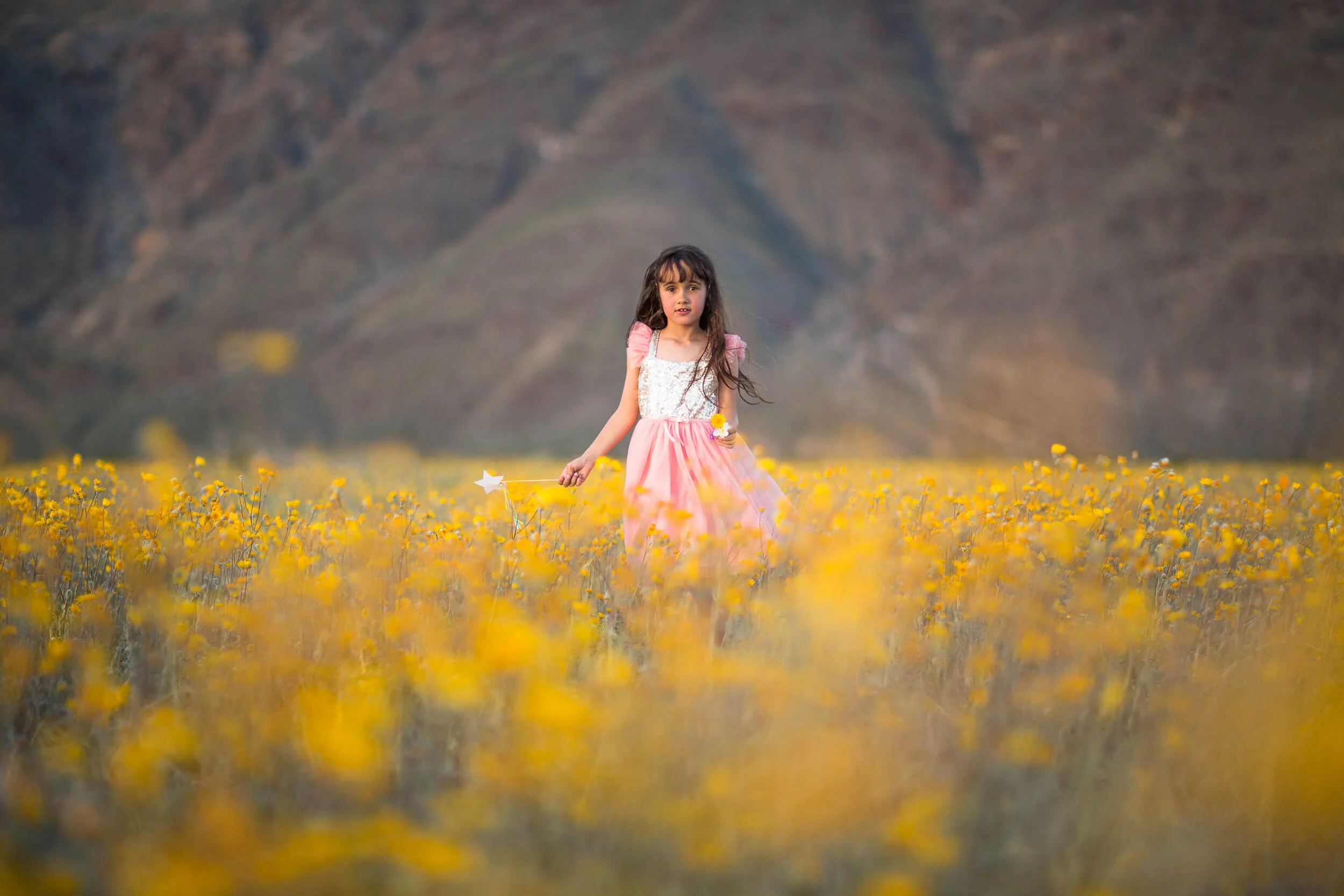 Young girl in a pink dress holding a fairy wand, walking through a field of yellow flowers with mountains in the background.