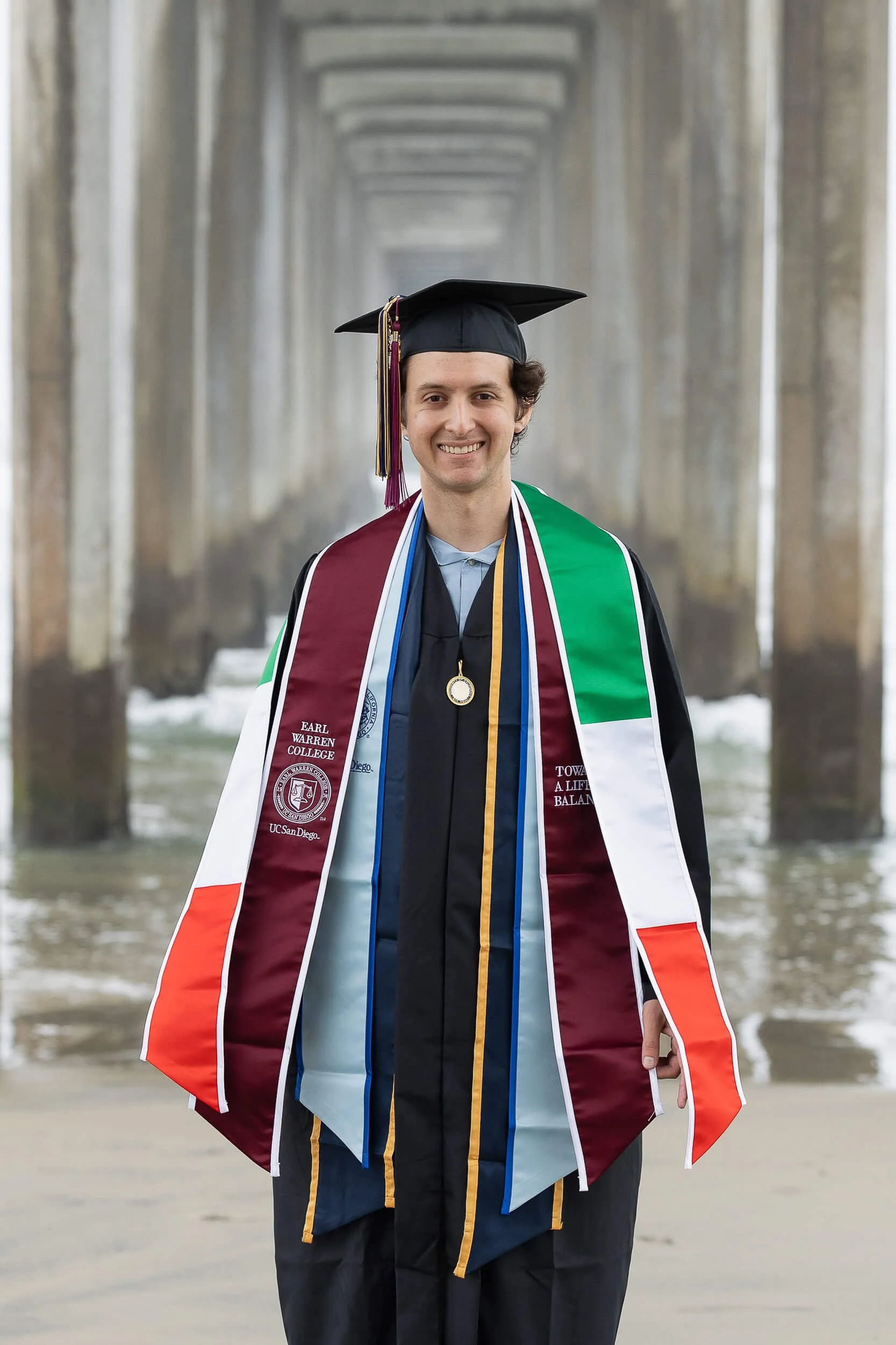 A young man in graduation cap and gown standing on a beach with a pier in the background.