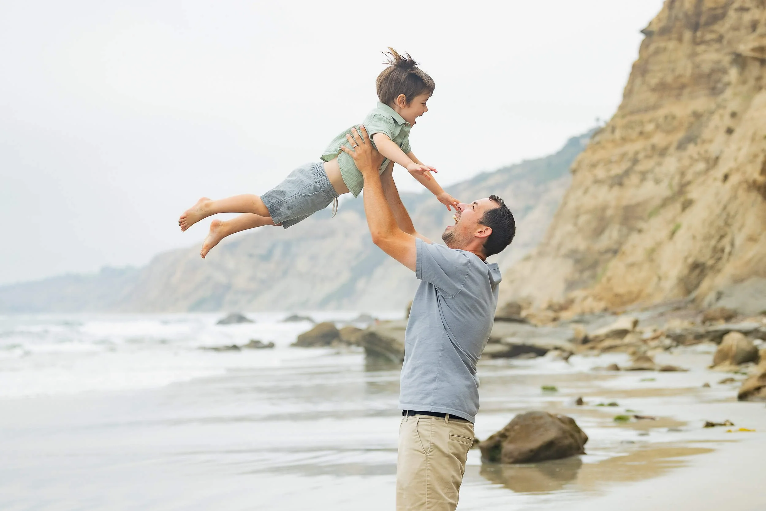 A man lifting a young boy by the beach, both smiling and reaching towards each other, with rocky cliffs and the ocean in the background.