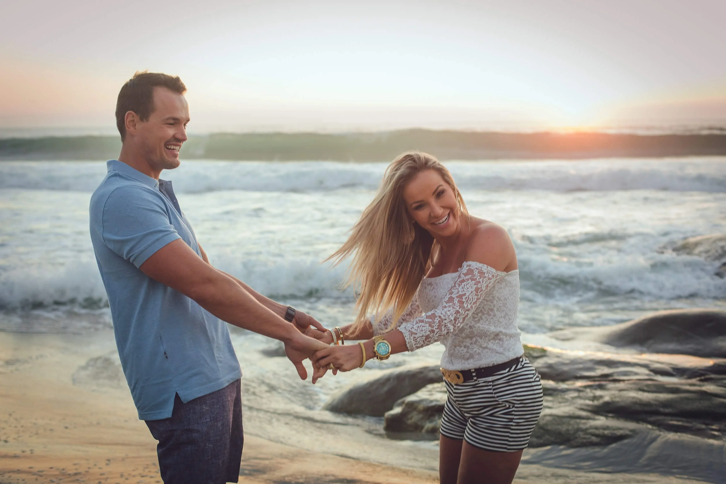 A smiling couple holding hands on the beach during sunset, with waves in the background.