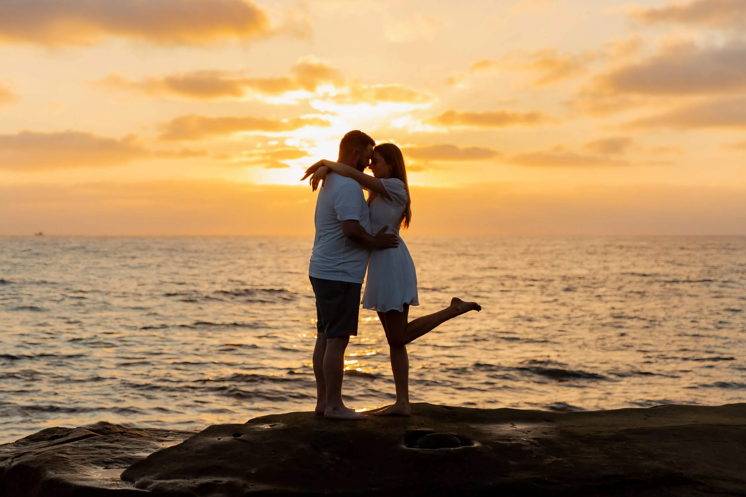 A couple hugging and kissing on a beach during sunset, with the woman lifting one leg in the air.