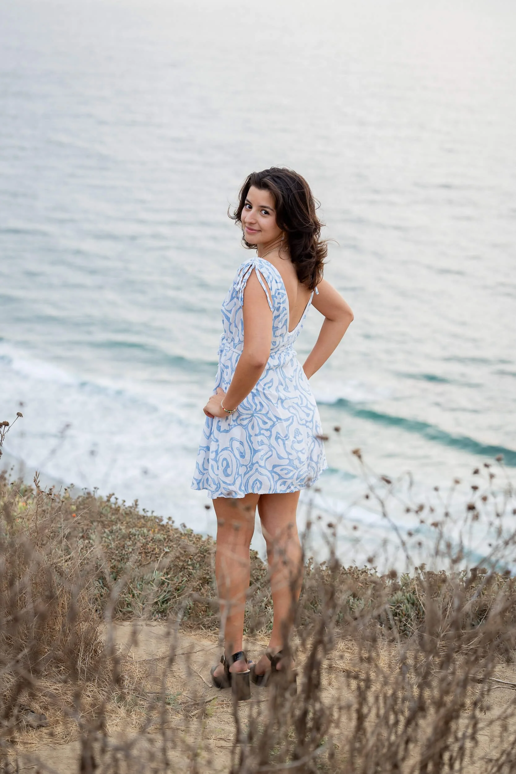 A woman in a blue and white dress walking on a sandy and grassy path near the ocean, turning back and smiling at the camera, with water and waves in the background.
