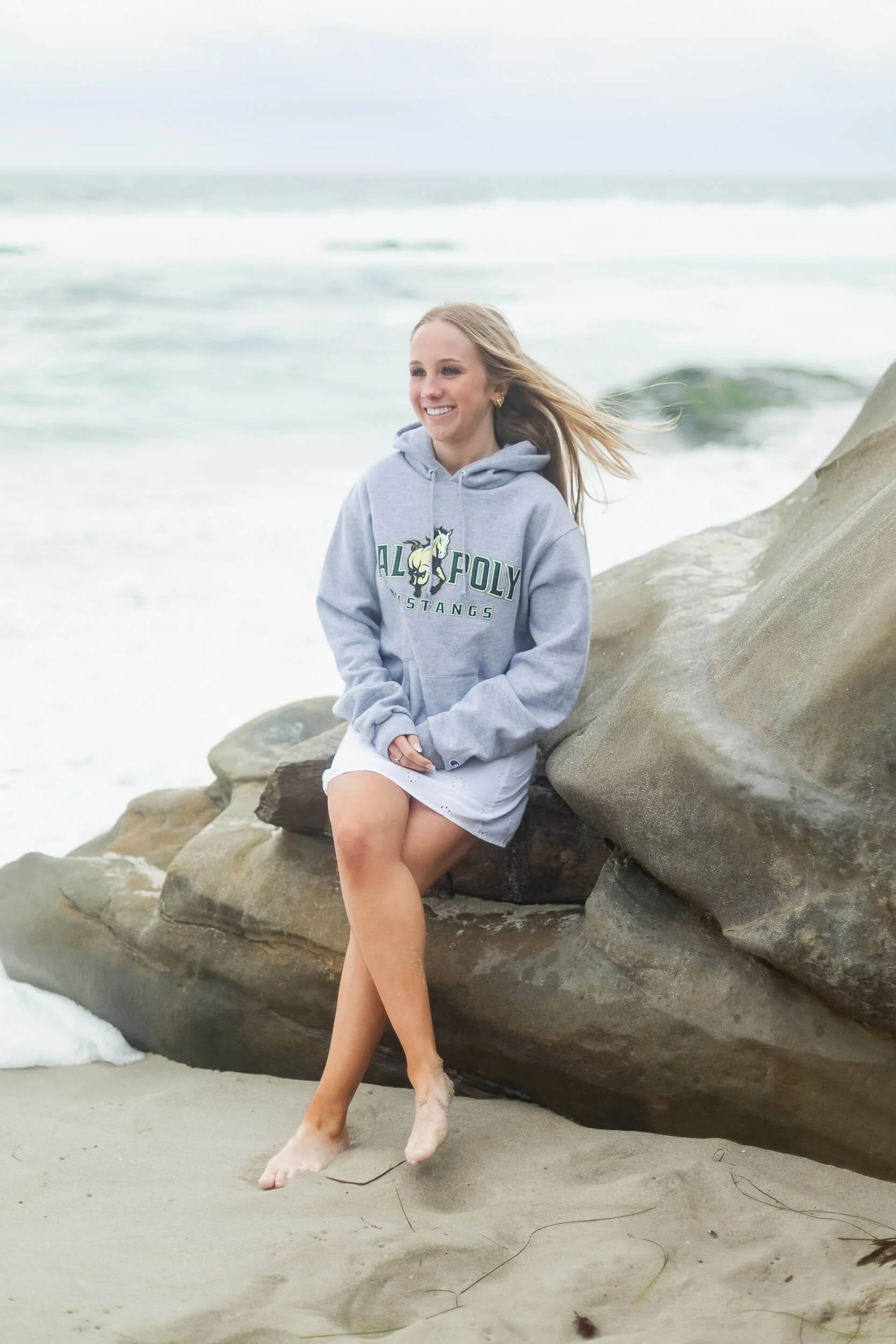 A young woman with blonde hair sitting on rocks on a beach, smiling, wearing a gray sweatshirt with 'Cal Poly Stangs' on it, and a white skirt, with waves behind her.